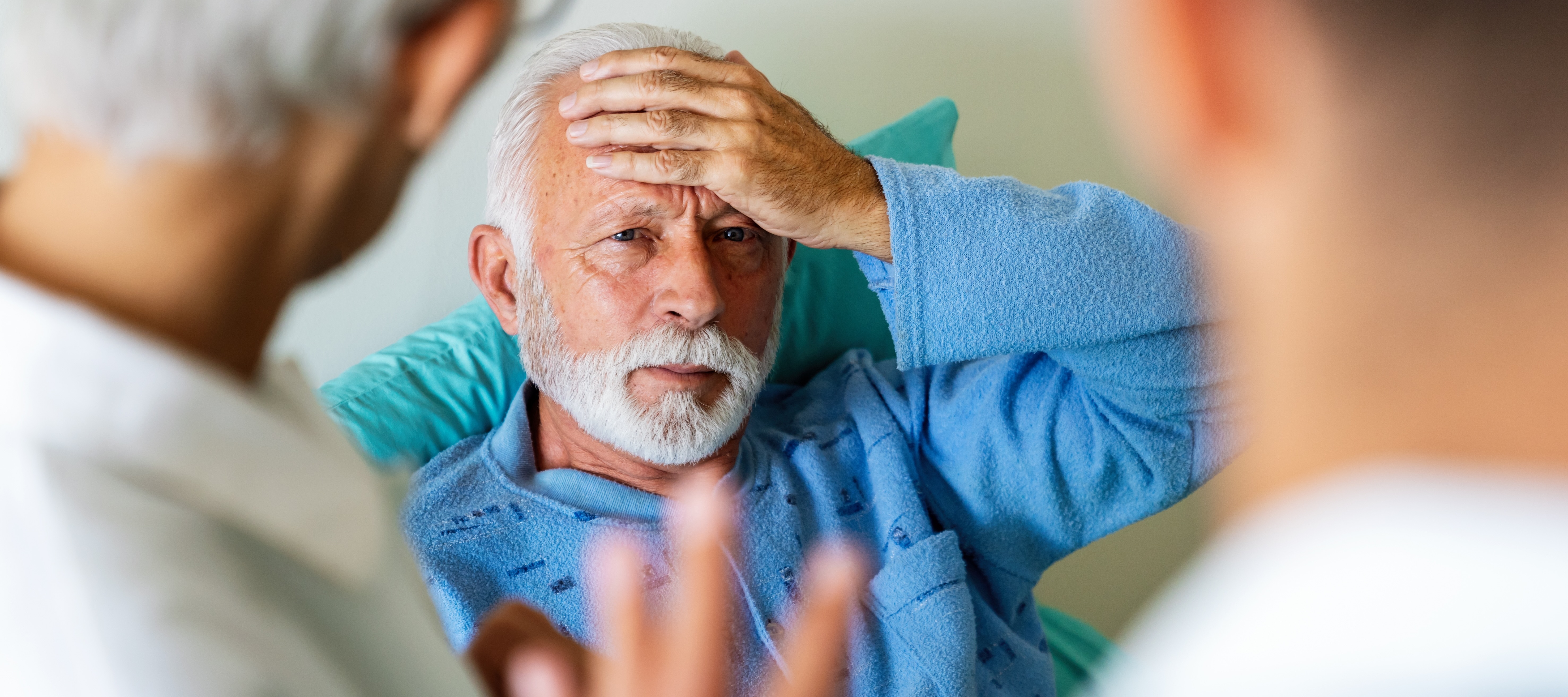 Gentleman with white hair holding his head in a healthcare setting with two healthcare workers looking on.