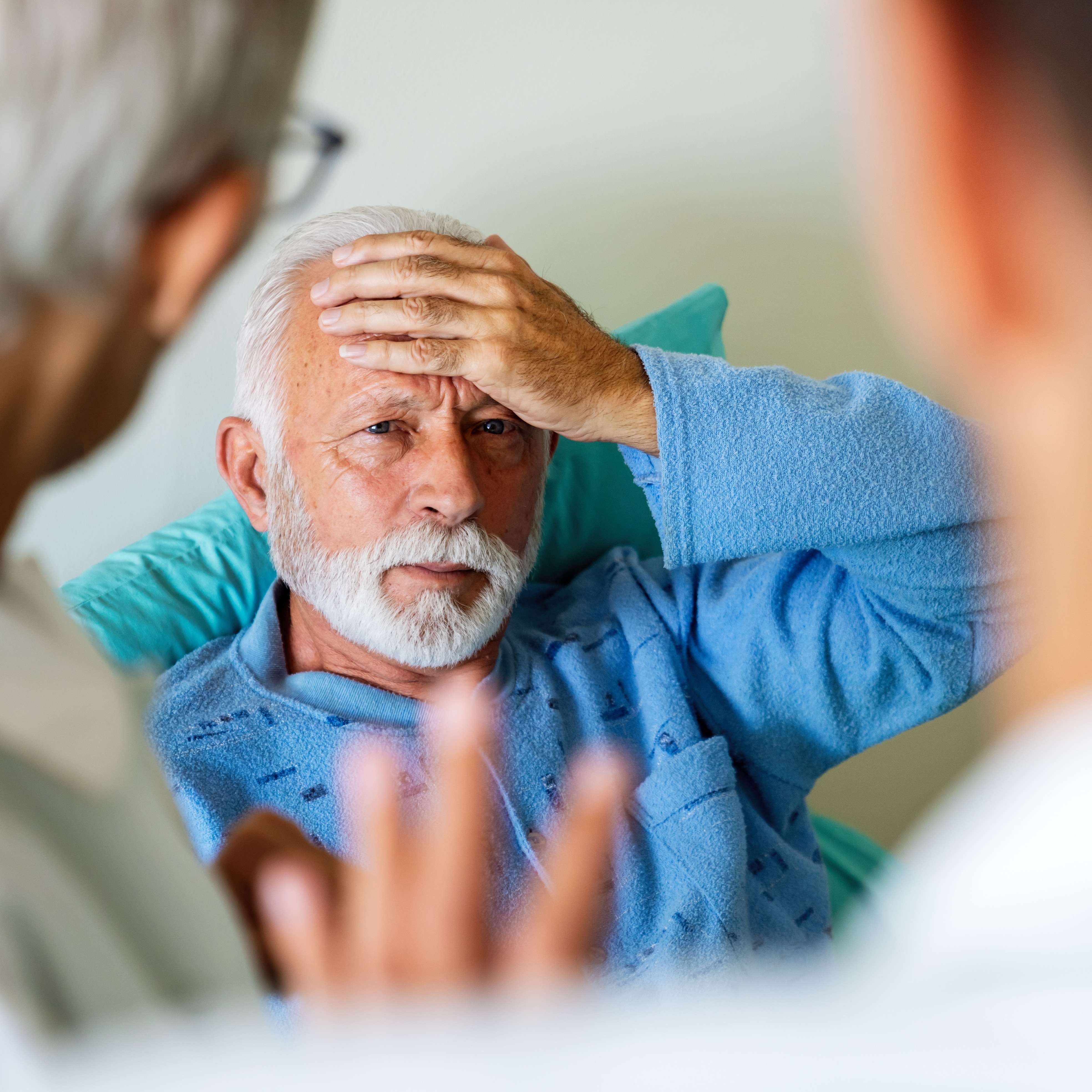 Gentleman with white hair holding his head in a healthcare setting with two healthcare workers looking on.