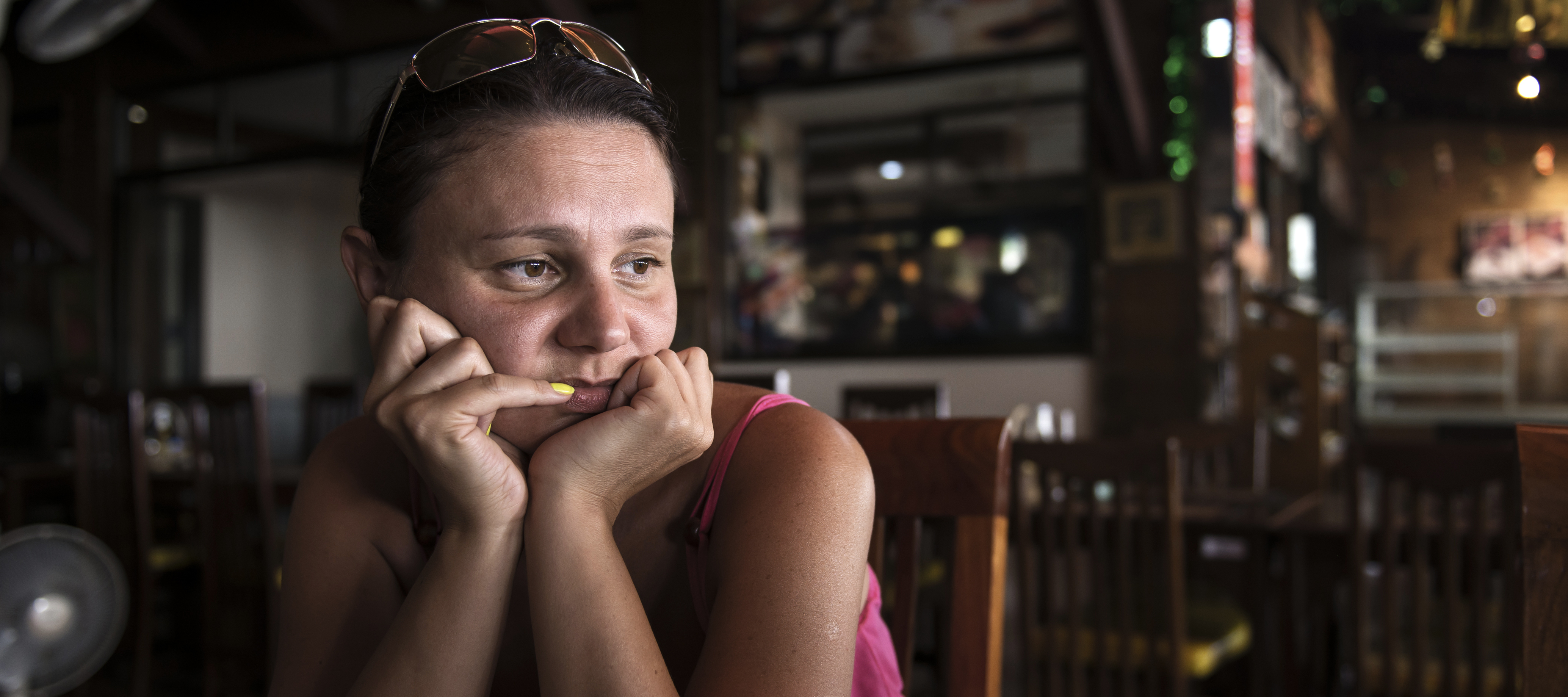 A woman sits in silent contemplation in a dim cafe, peering over yellow-painted nails.