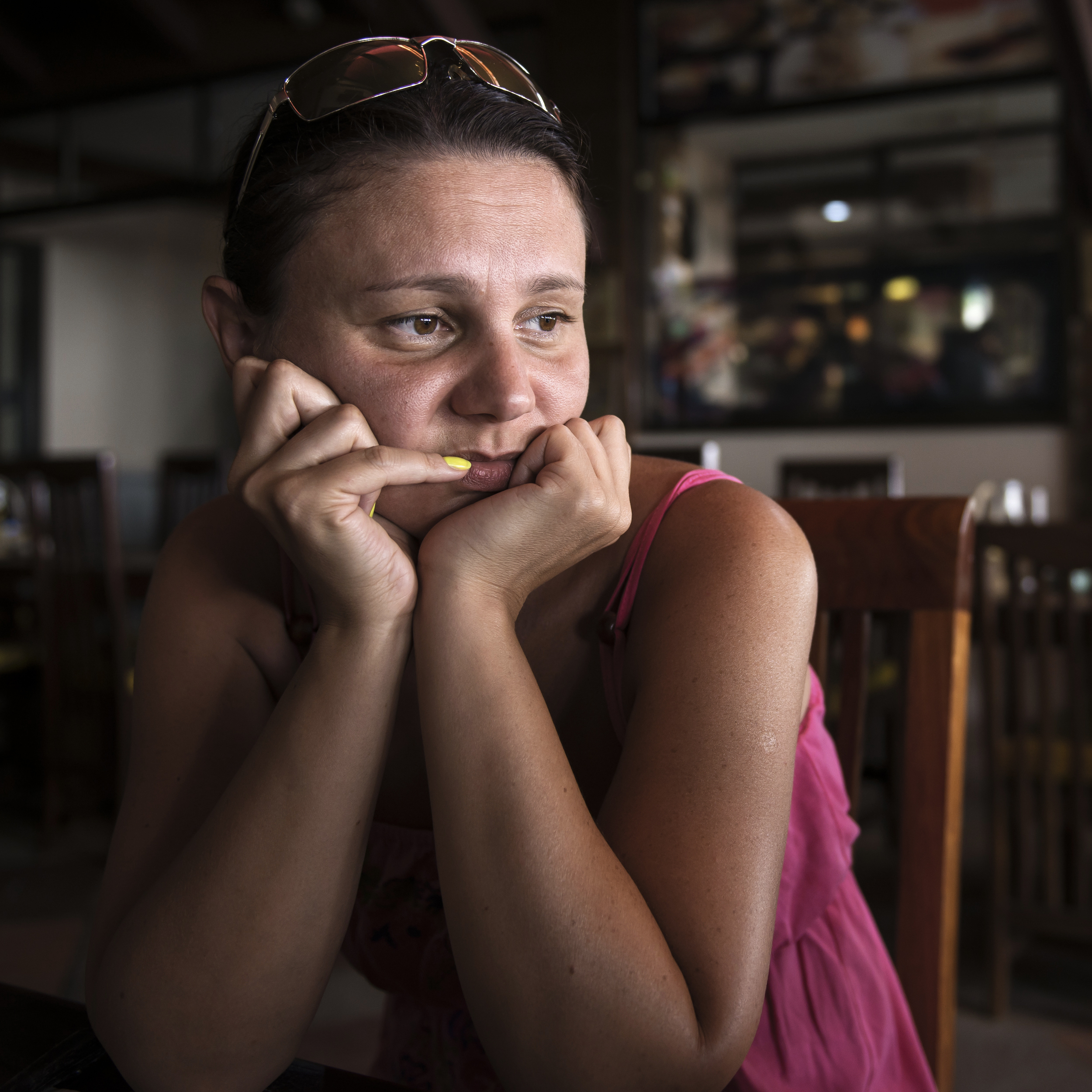 A woman sits in silent contemplation in a dim cafe, peering over yellow-painted nails.