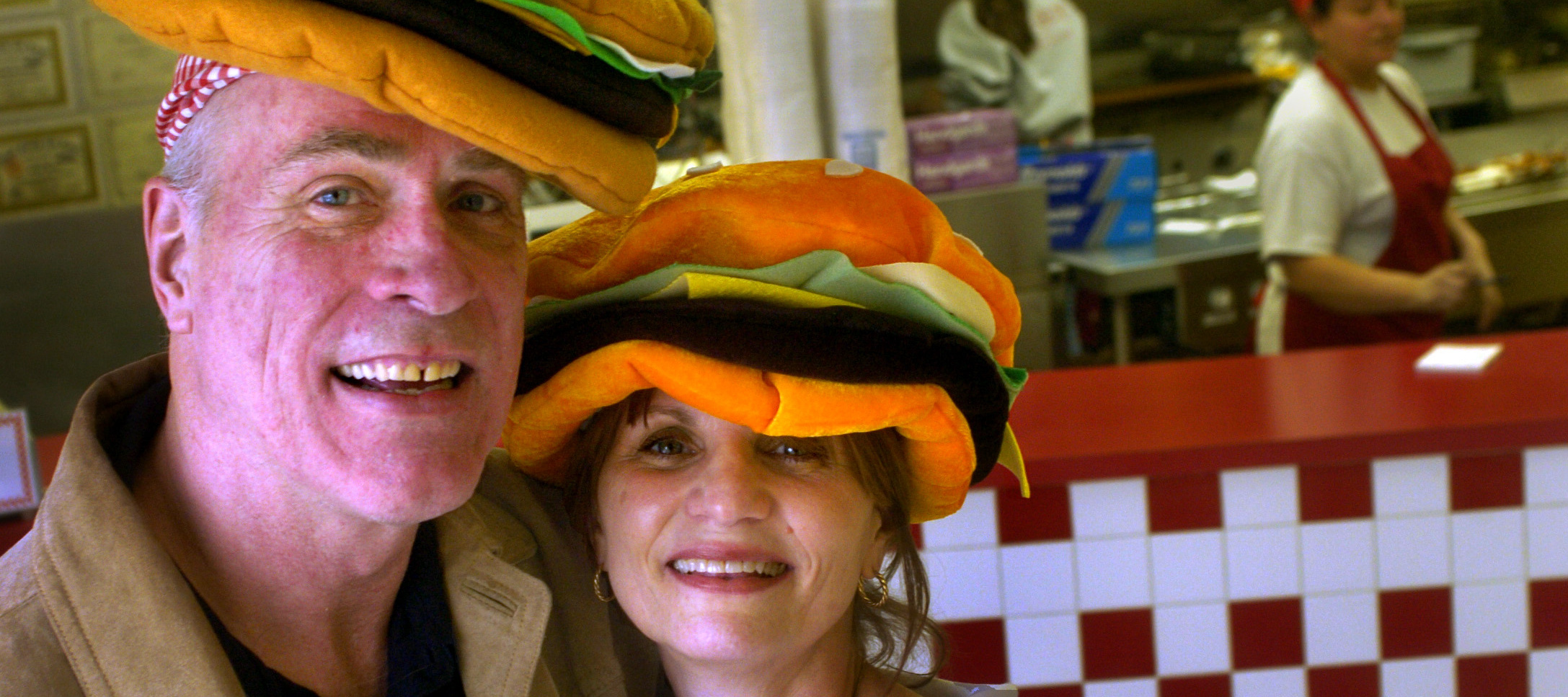 Jerry and Janie Murrell smile while wearing hamburger shaped hates inside of a Five Guys restaurant.