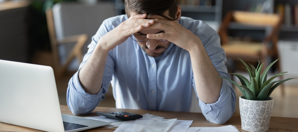 Frustrated man sits at desk in despair, checking money documents, overwhelmed by finances.