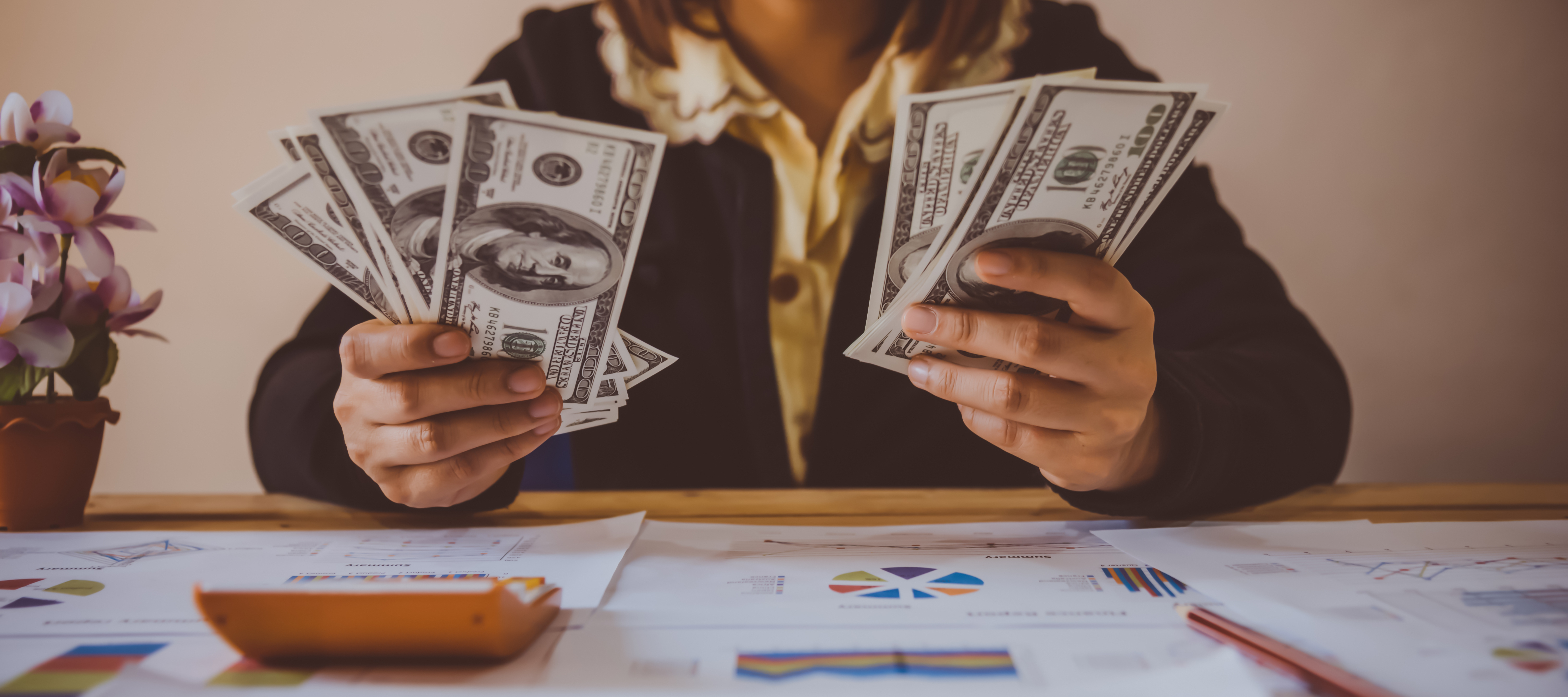 Close up of a woman holding hundred dollar bills in her hand while staring at investing reports on her desk.