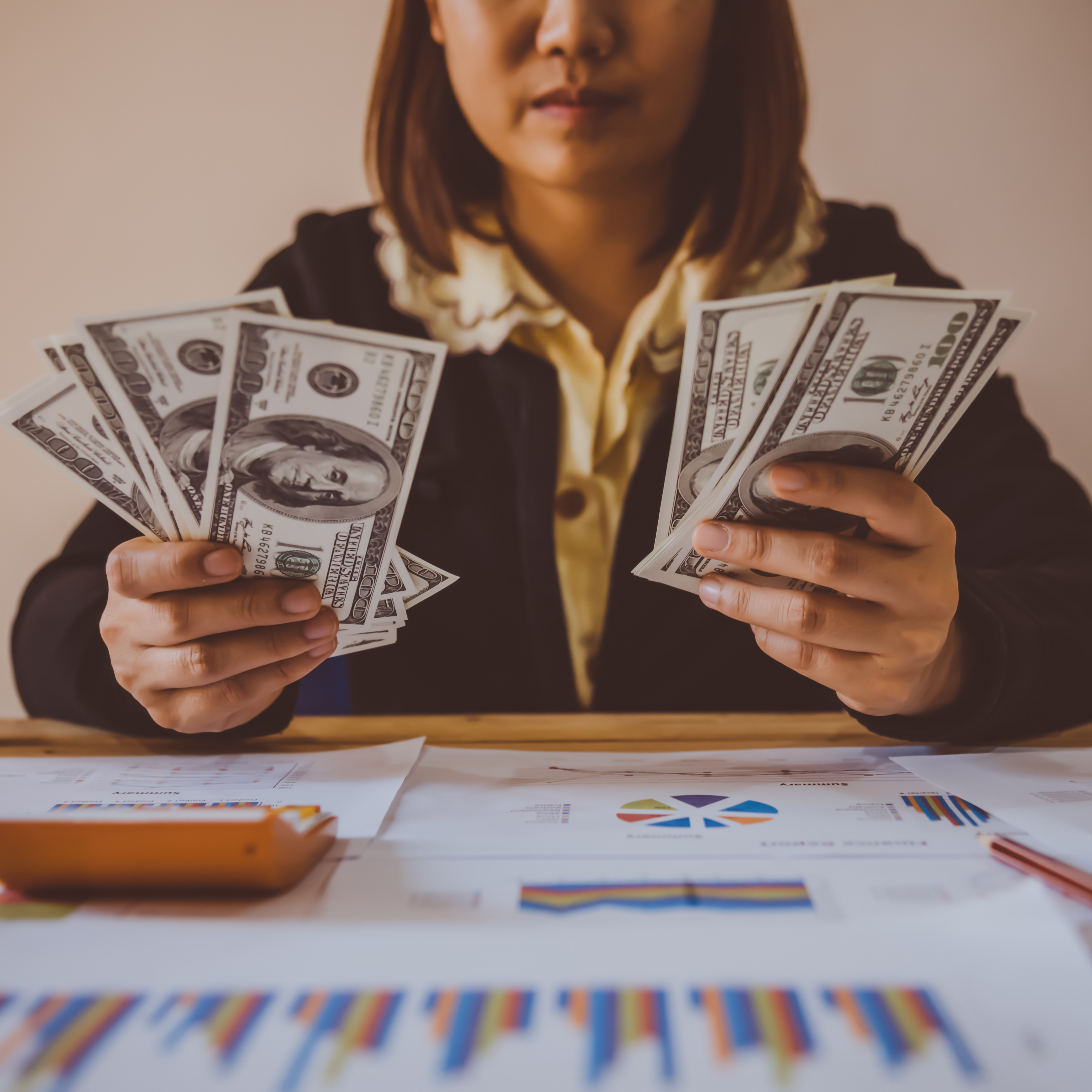 Close up of a woman holding hundred dollar bills in her hand while staring at investing reports on her desk.