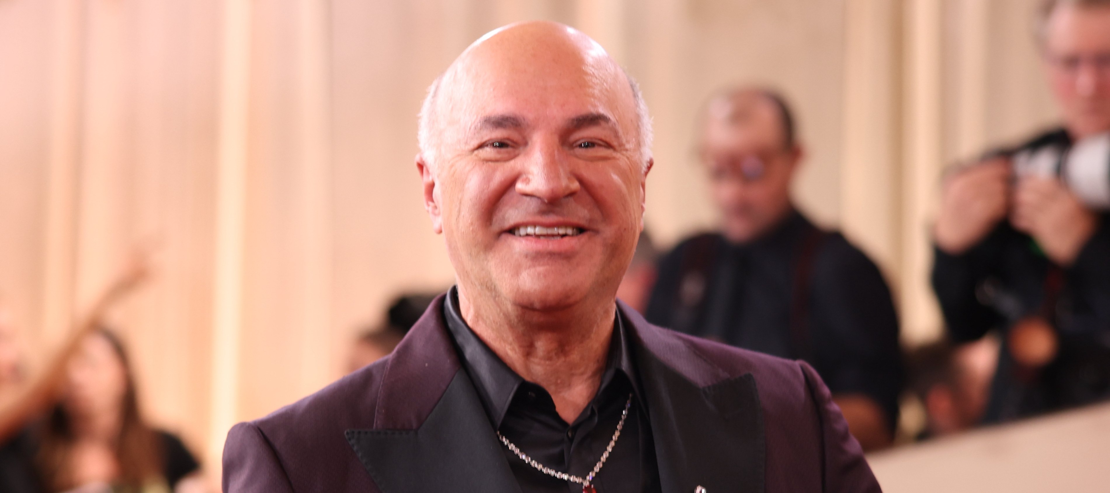 Shark Tank star Kevin O'Leary spreads his arms wide and smiles at the camera in an immaculate purple and black suit.