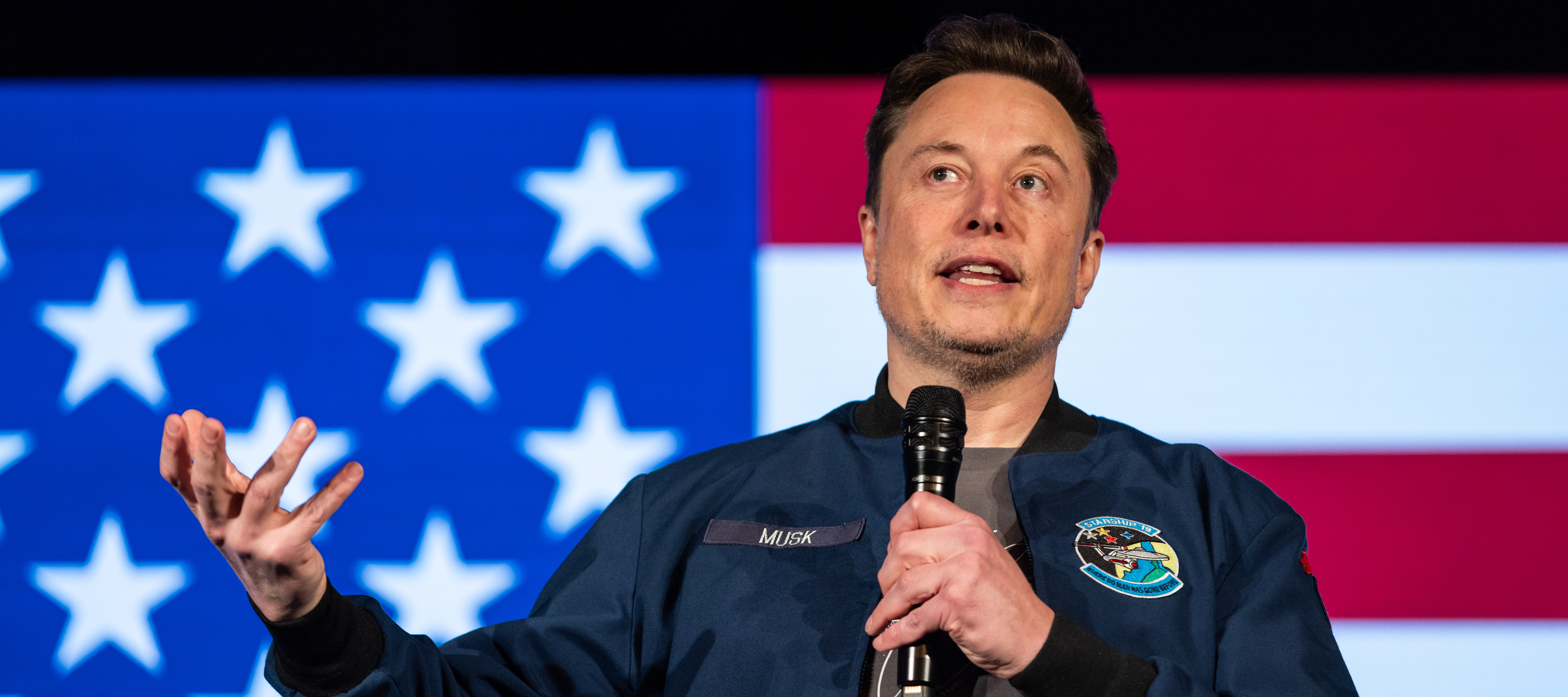Elon musk stands in front of the American flag, gesturing outwards with one hand and looking upwards.