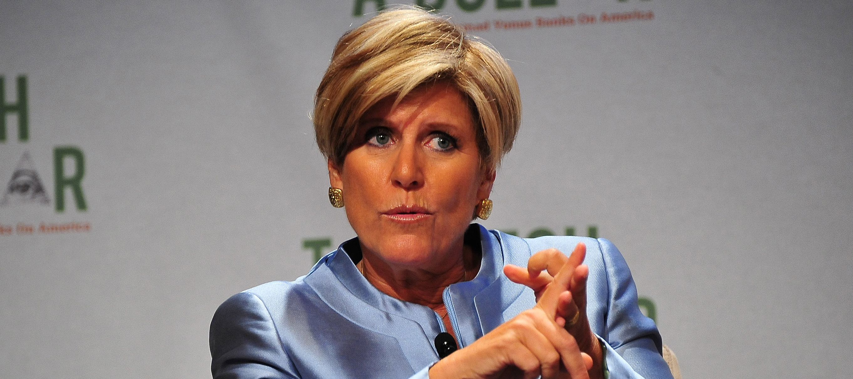 Suze Orman sits on a chair onstage with her hands folded together, on the verge of speaking.