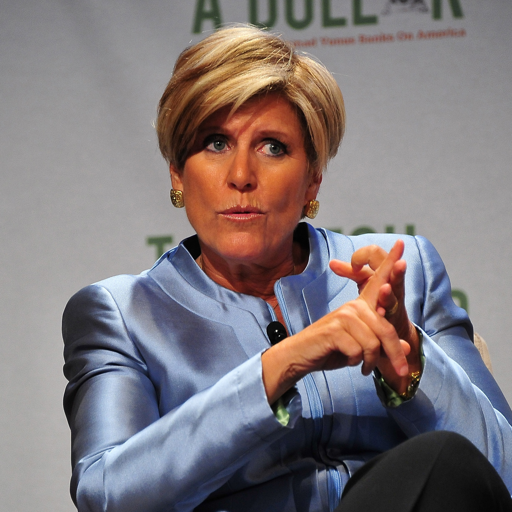 Suze Orman sits on a chair onstage with her hands folded together, on the verge of speaking.