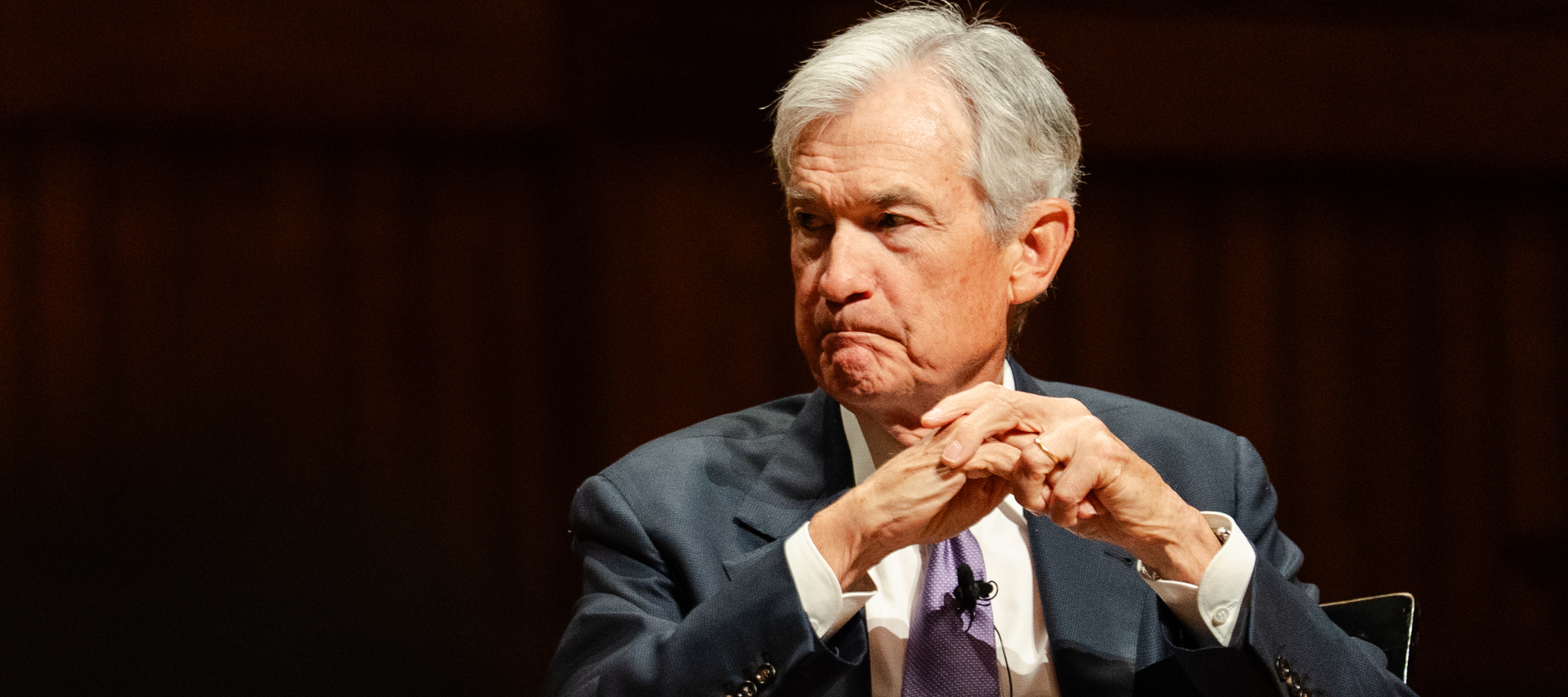 Jerome Powell sits quietly on a dark stage, hands folded in front of his face and looking askew into the middle distance.