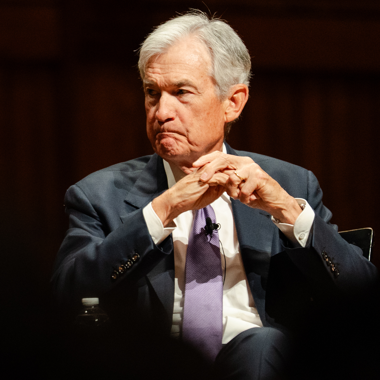 Jerome Powell sits quietly on a dark stage, hands folded in front of his face and looking askew into the middle distance.