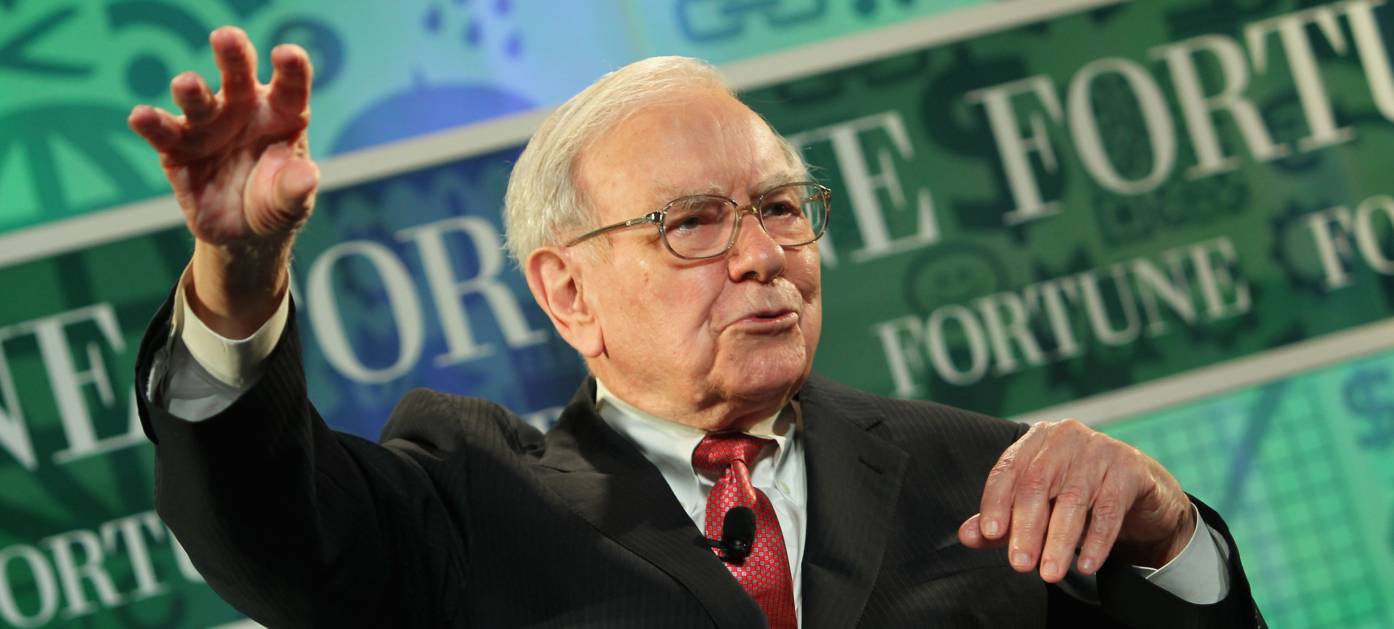Warren Buffett holds up one hand to the sky while speaking against a green backdrop.