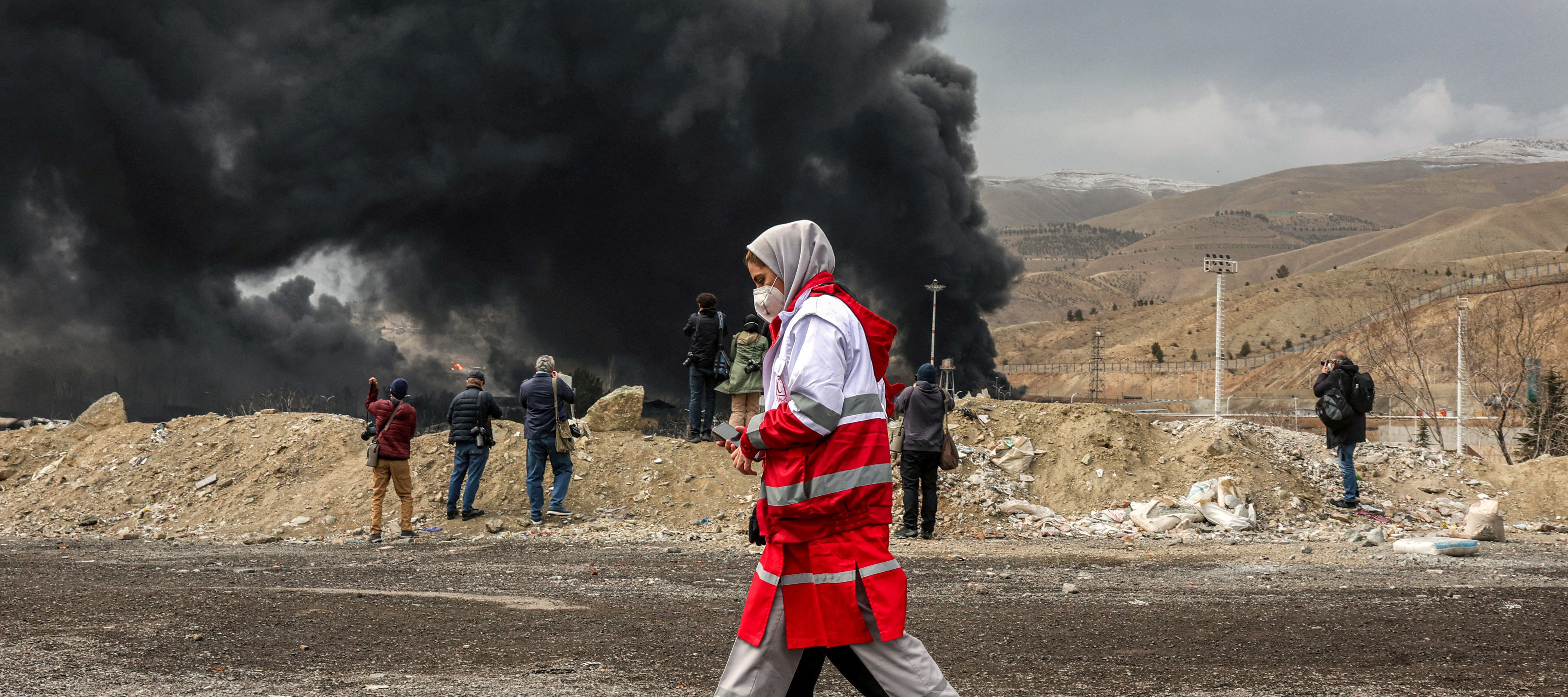Members of Iran's Red Crescent society walk near smoke plumes from a fire following an overnight airstrike on the Shahran oil refinery in northwestern Tehran on March 8, 2026.