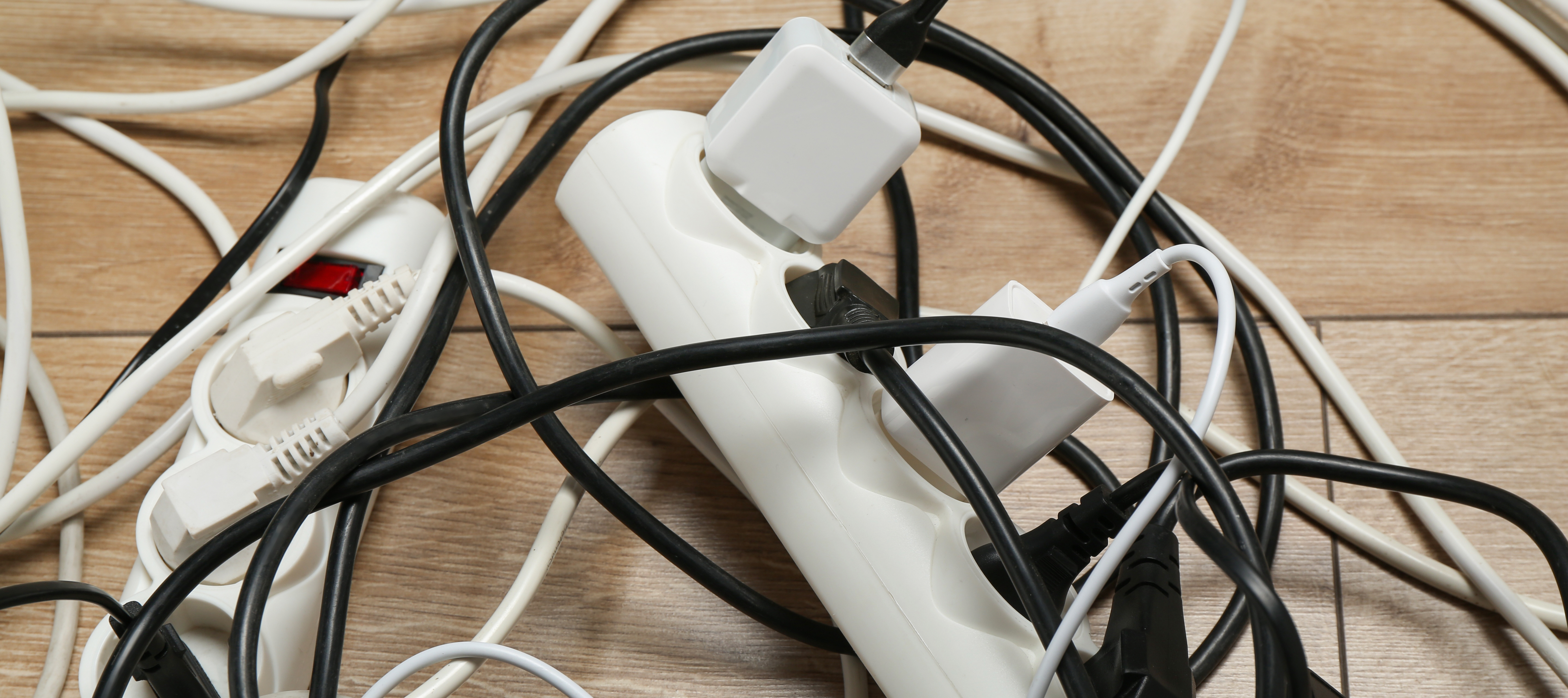 An overhead shot shows a chaotic arrangement of white and black electrical cords and white extension plugs resting on a light brown wooden floor.