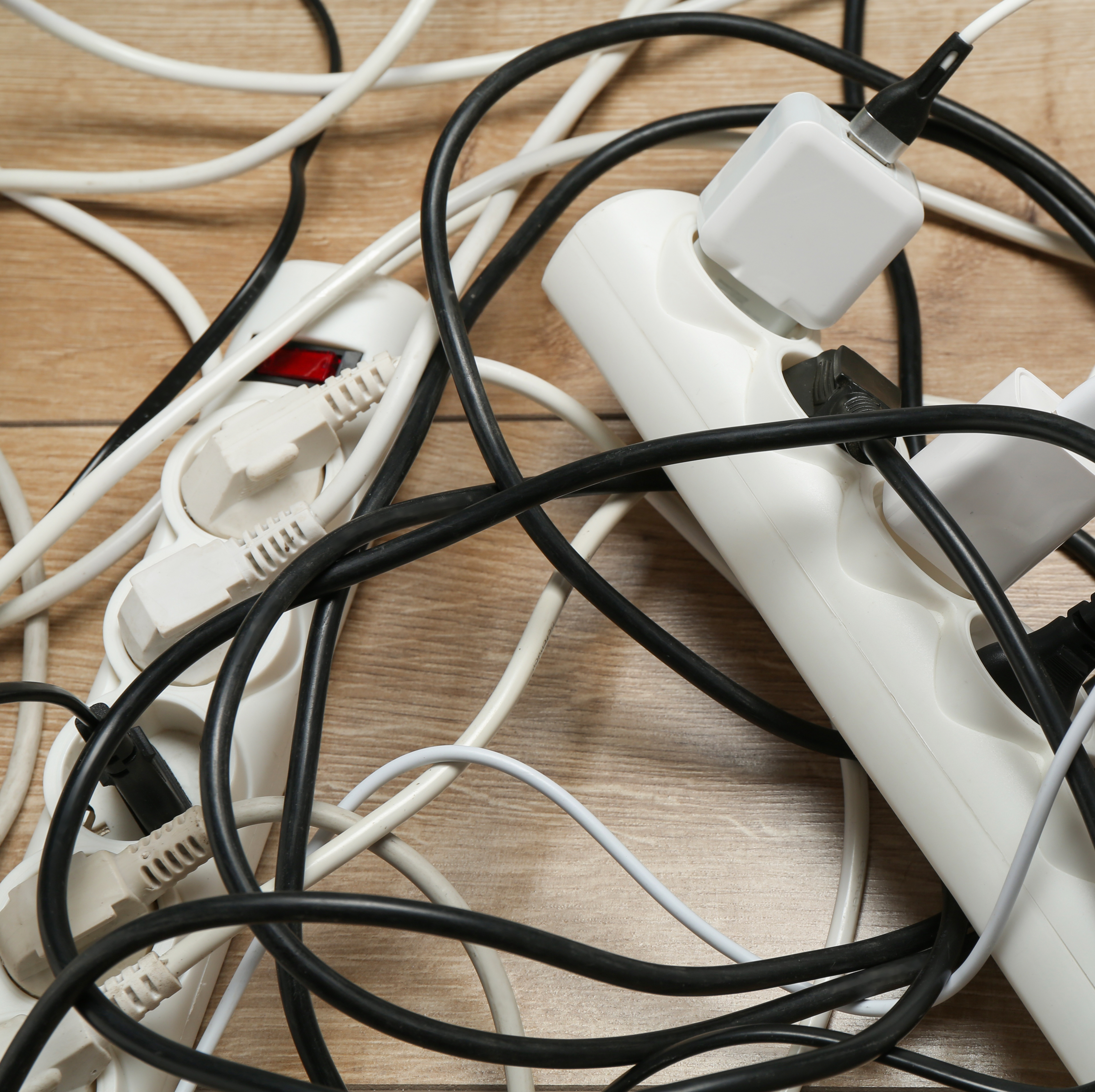 An overhead shot shows a chaotic arrangement of white and black electrical cords and white extension plugs resting on a light brown wooden floor.