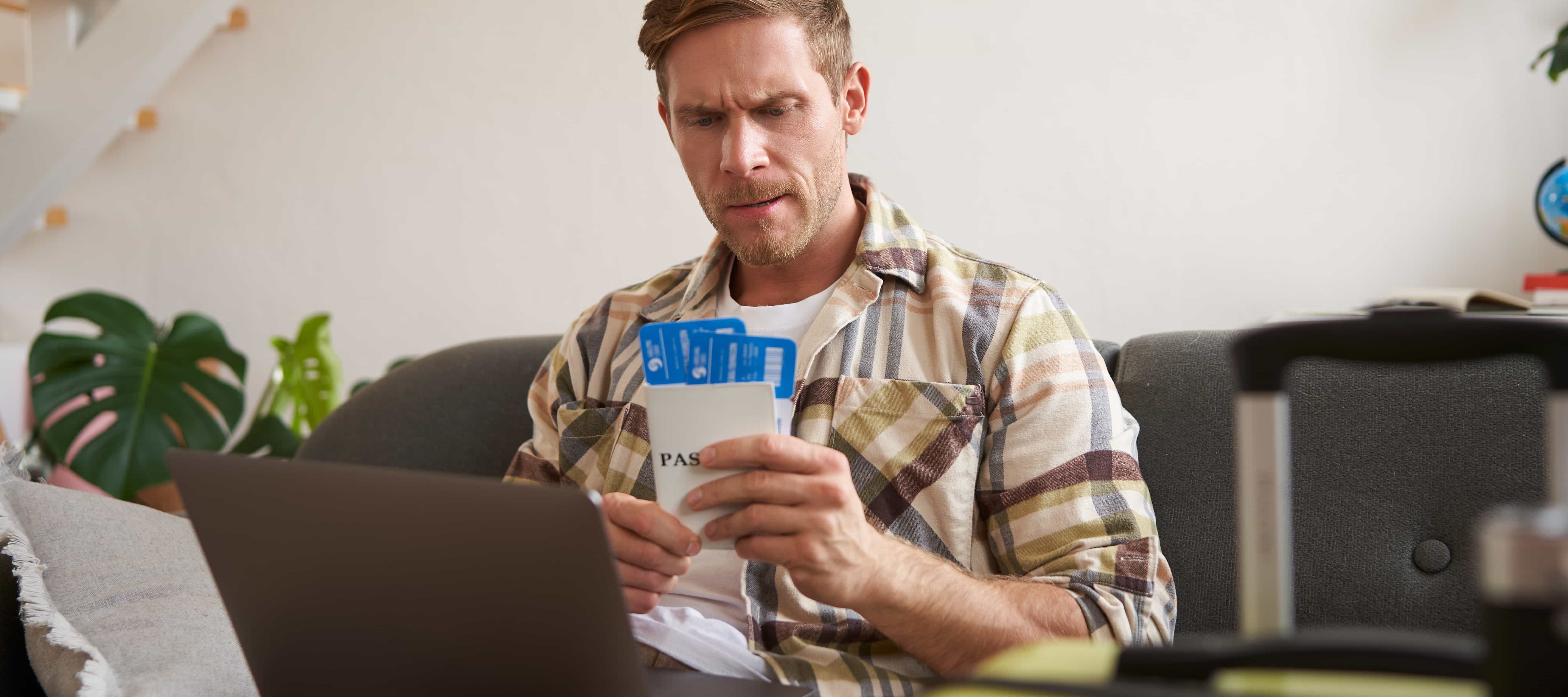 A photo of a man using his laptop and holding flight tickets beside his suitcase