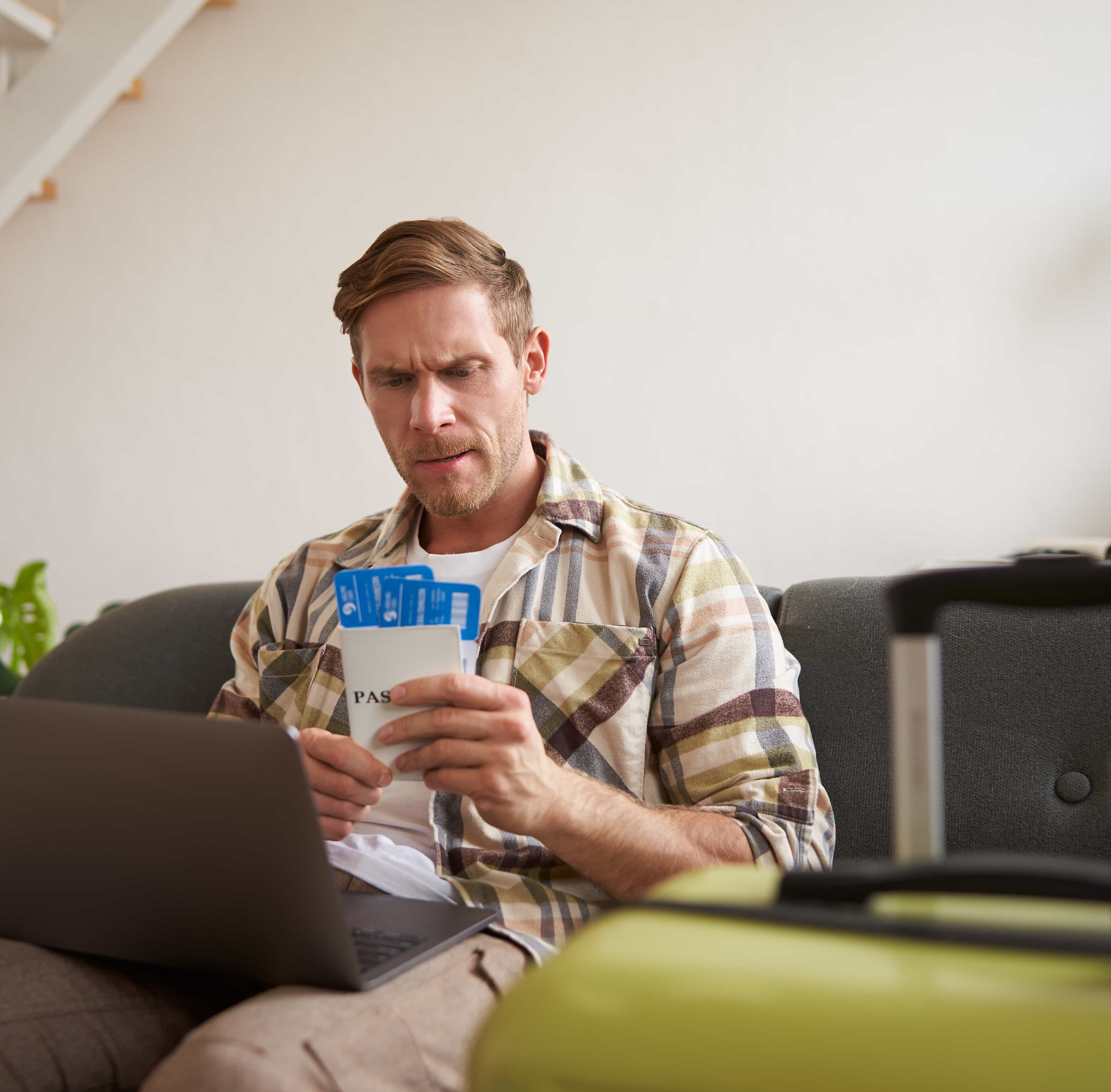 A photo of a man using his laptop and holding flight tickets beside his suitcase