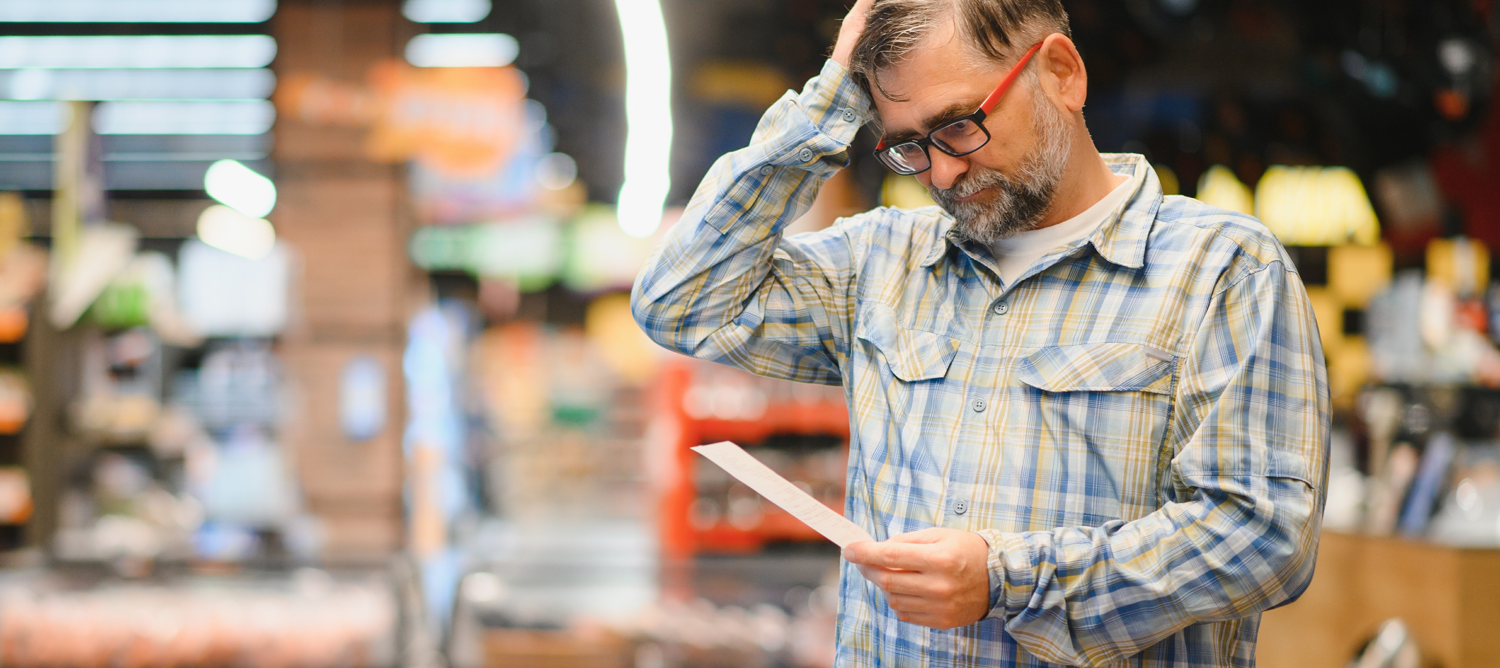 Middle-aged man looks at a grocery list or receipt with stress on his face inside of a store.