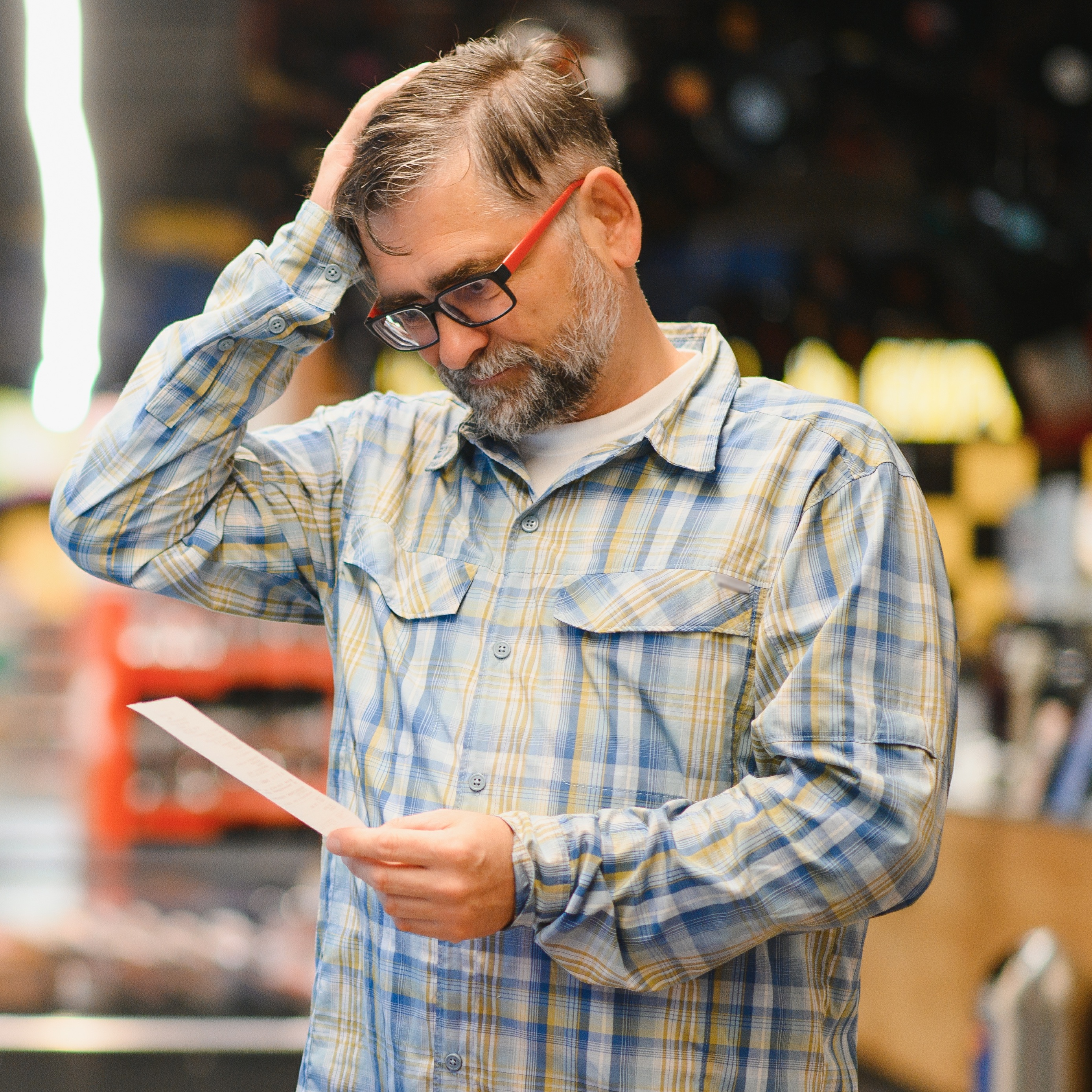 Middle-aged man looks at a grocery list or receipt with stress on his face inside of a store.