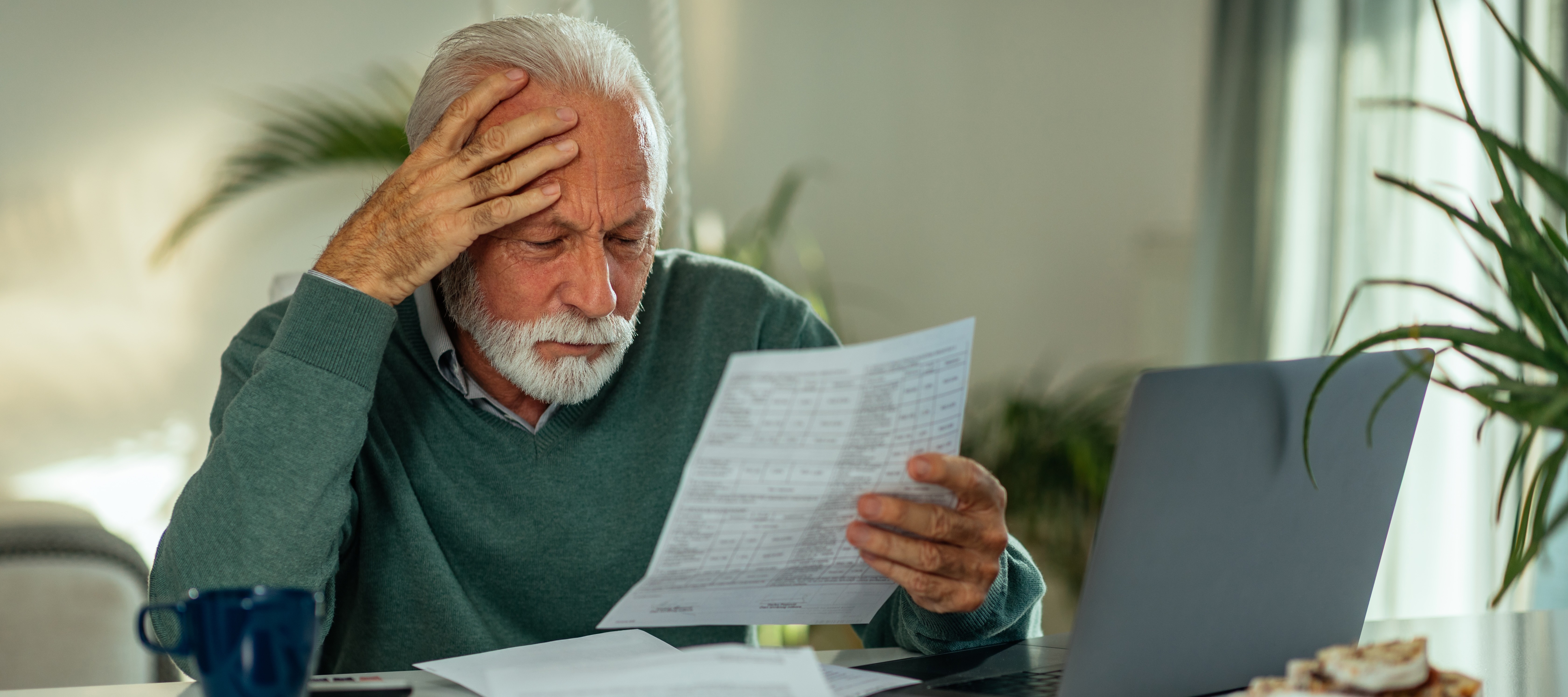 A senior man with grey hair and a white beard sits at a bright white table in his home, reviewing documents, looking worried.