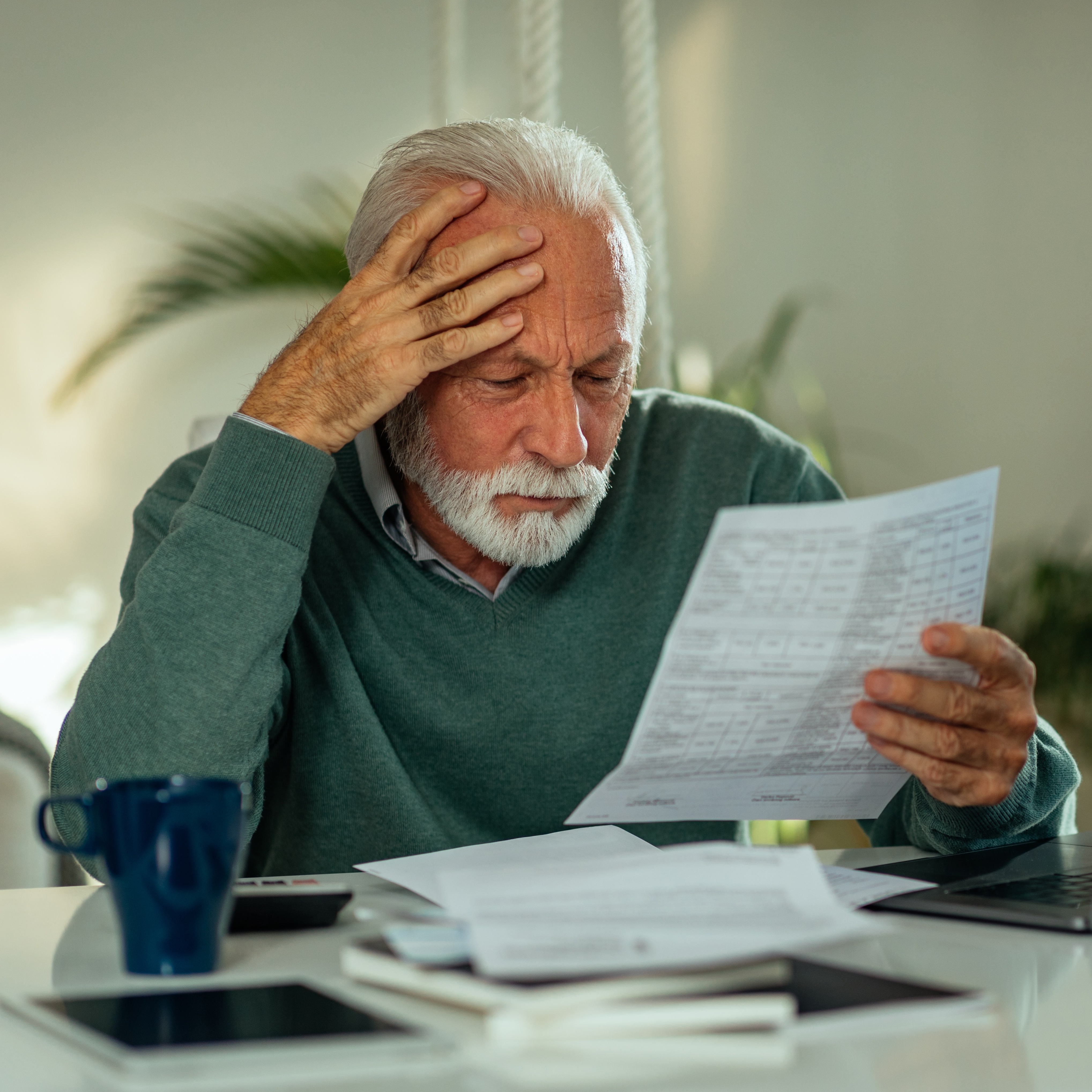A senior man with grey hair and a white beard sits at a bright white table in his home, reviewing documents, looking worried.