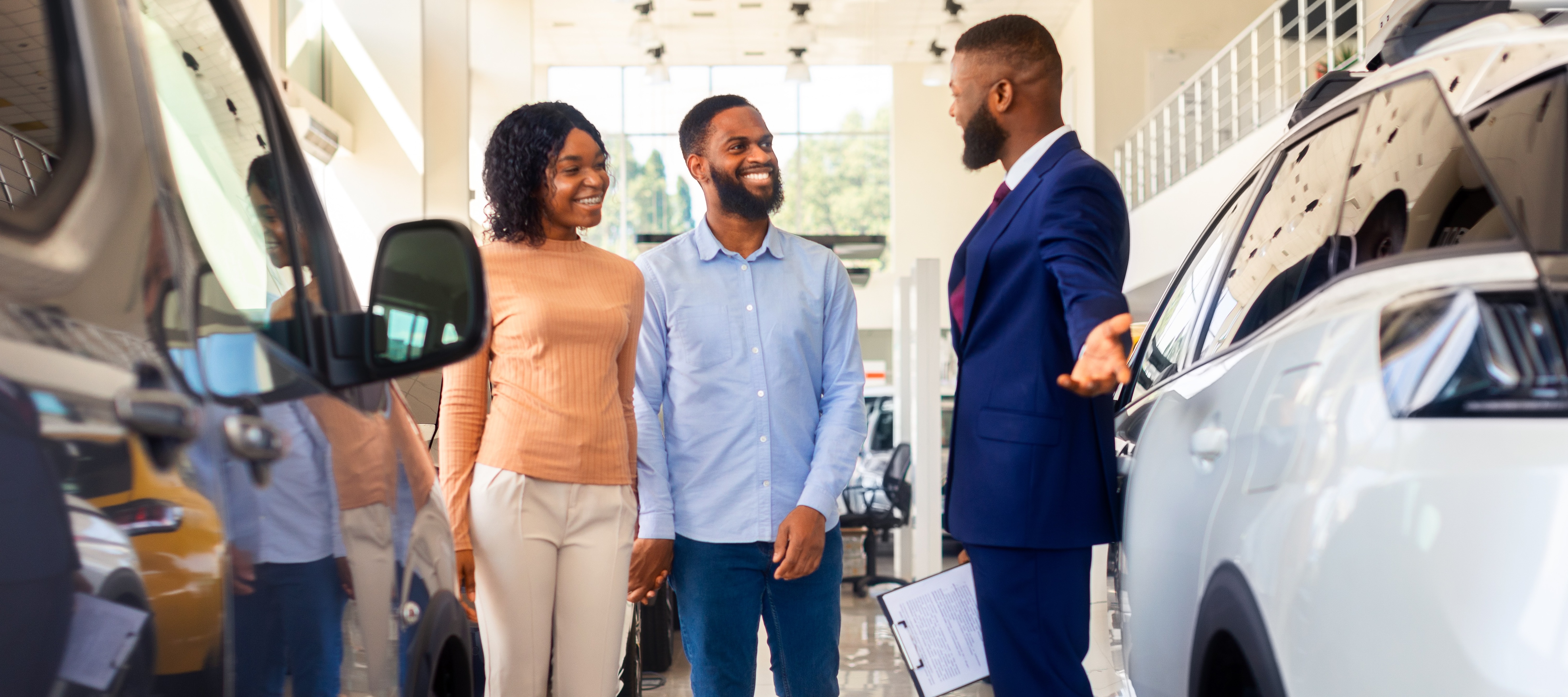 Car salesman gestures toward vehicles while shoppers stand nearby smiling in an indoor car lot.