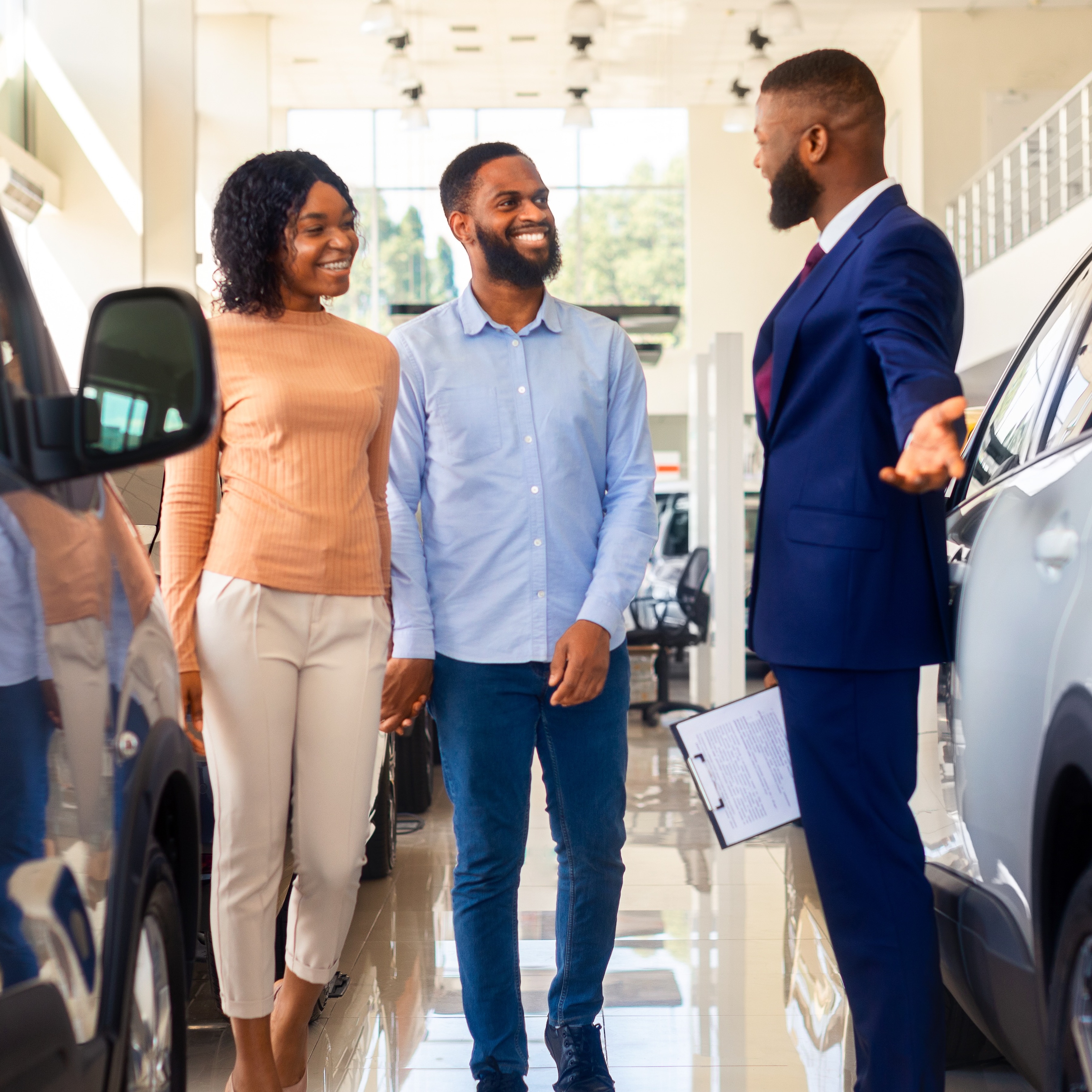 Car salesman gestures toward vehicles while shoppers stand nearby smiling in an indoor car lot.