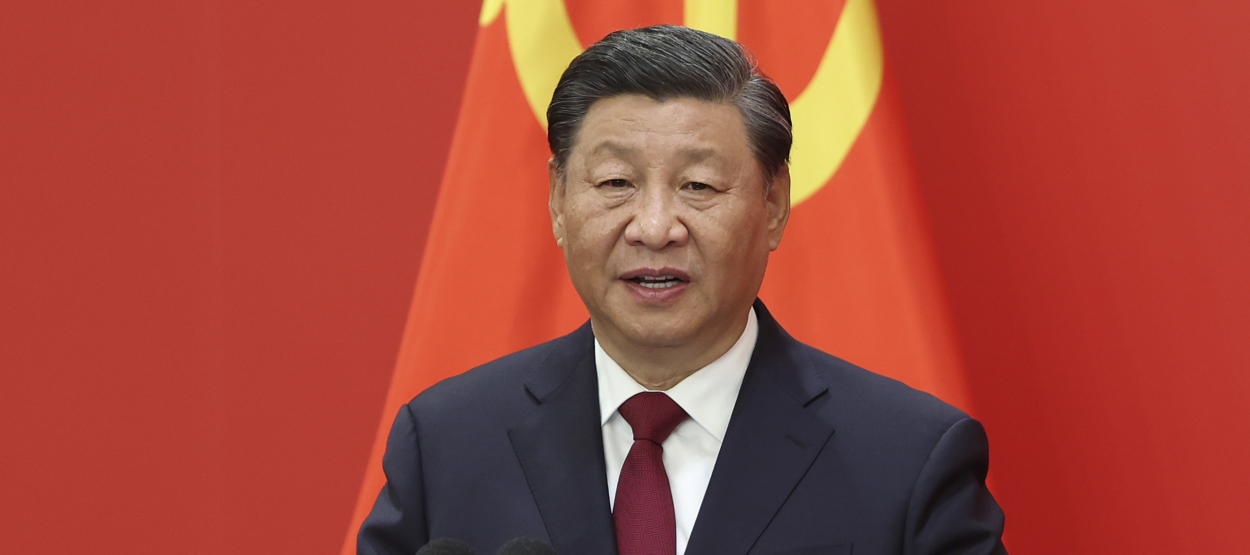 President Xi Jinping speaks at a podium against a stark red backdrop, featuring a hammer and sickle in yellow.