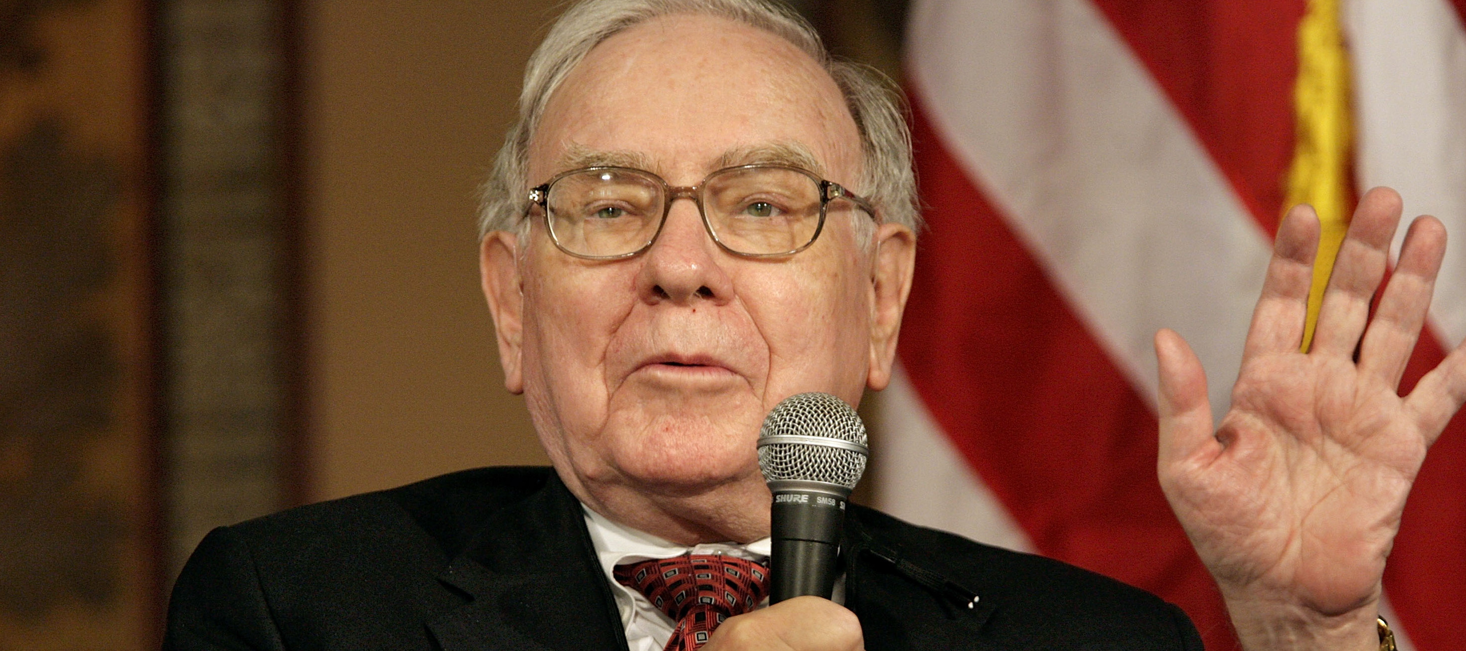 Warren Buffett raises one hand while speaking into a microphone.