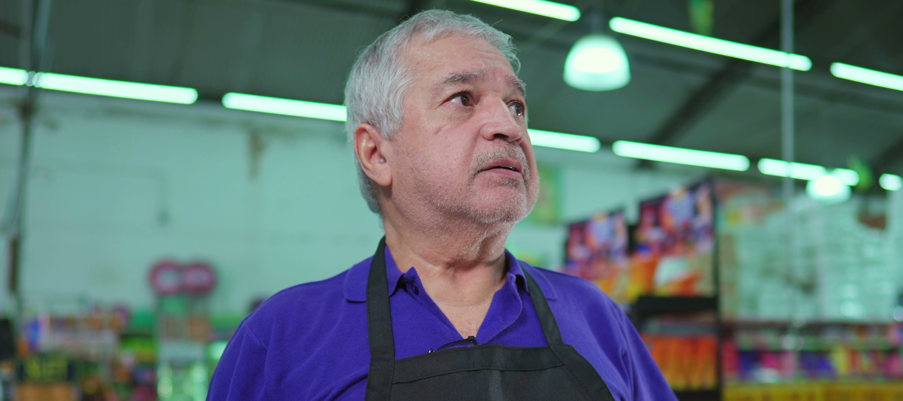 A worried grocery store manager stands in a dimly lit warehouse, looking up and away from a clipboard.