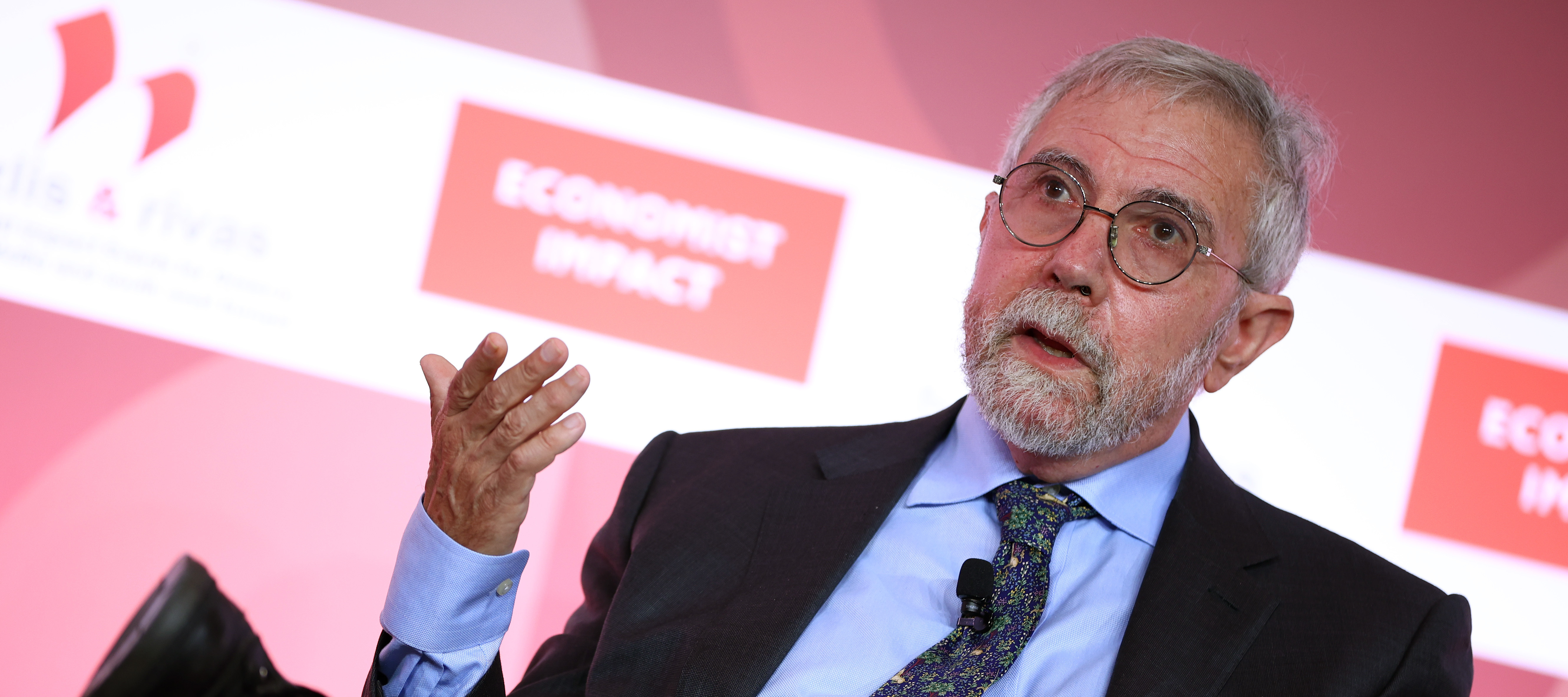 Paul Krugman speaks onstage seated in a chair with one hand raised.