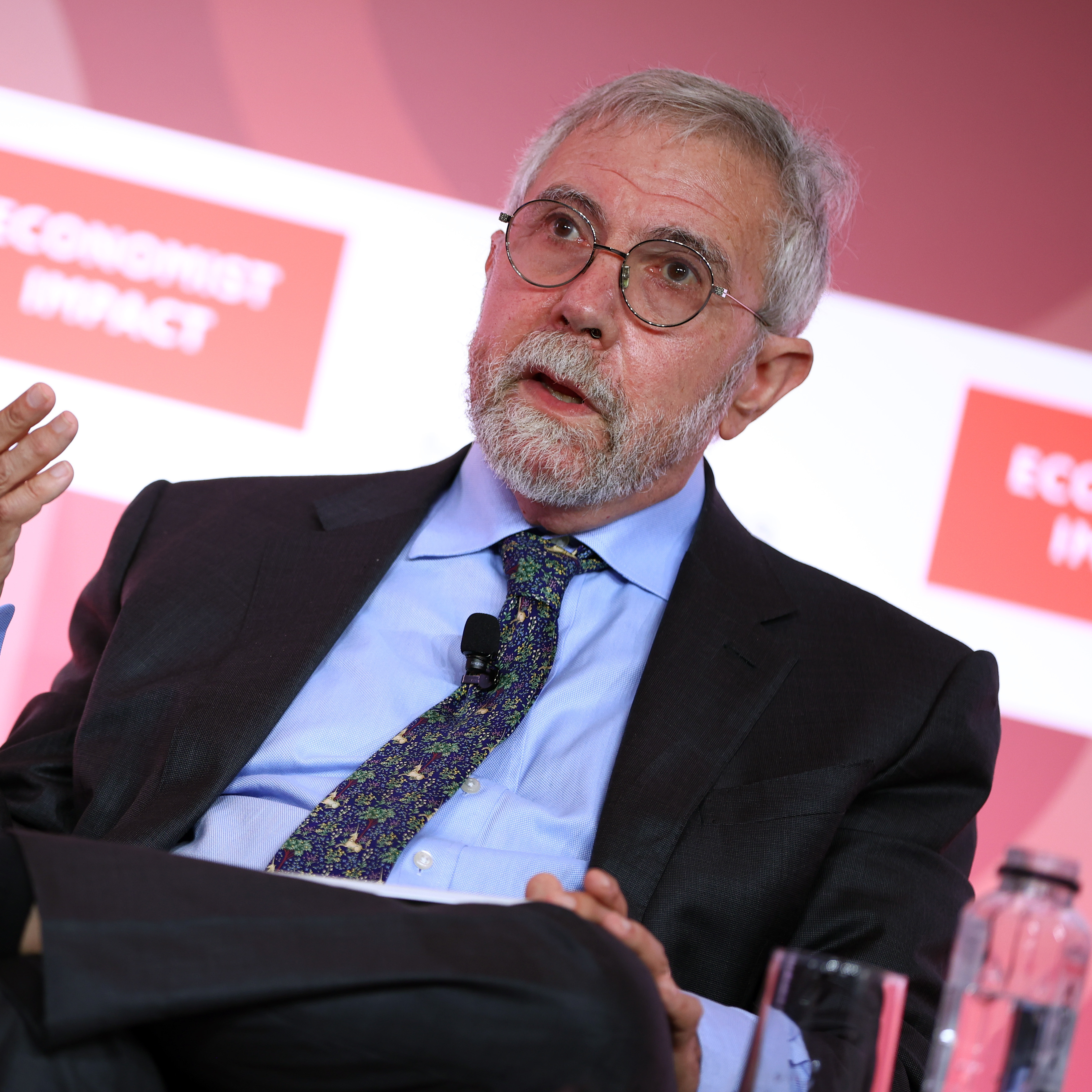 Paul Krugman speaks onstage seated in a chair with one hand raised.