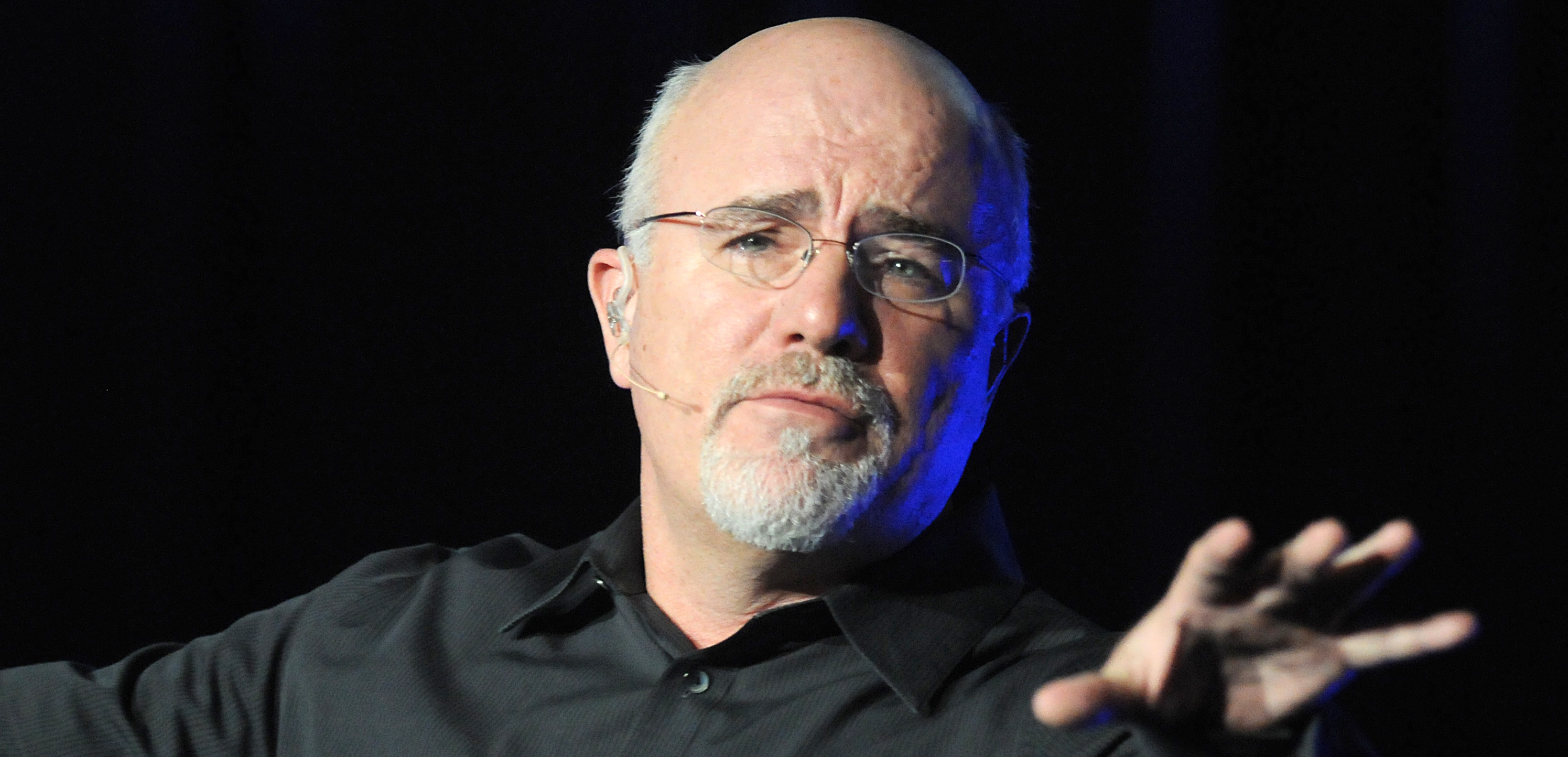 Dave Ramsey spreads out one hand on stage and looks directly at the camera.