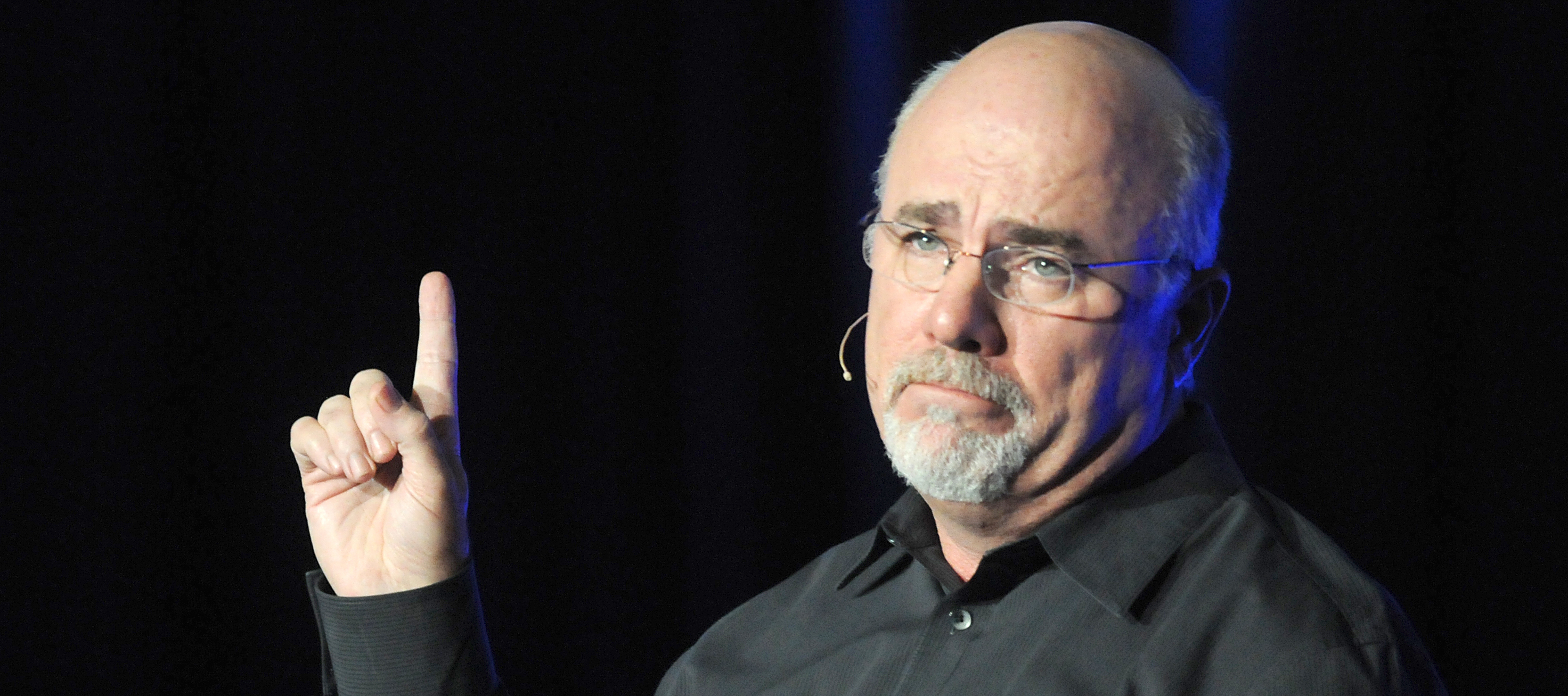 Dave Ramsey stands on stage looking solemn and holding a single finger to the sky.