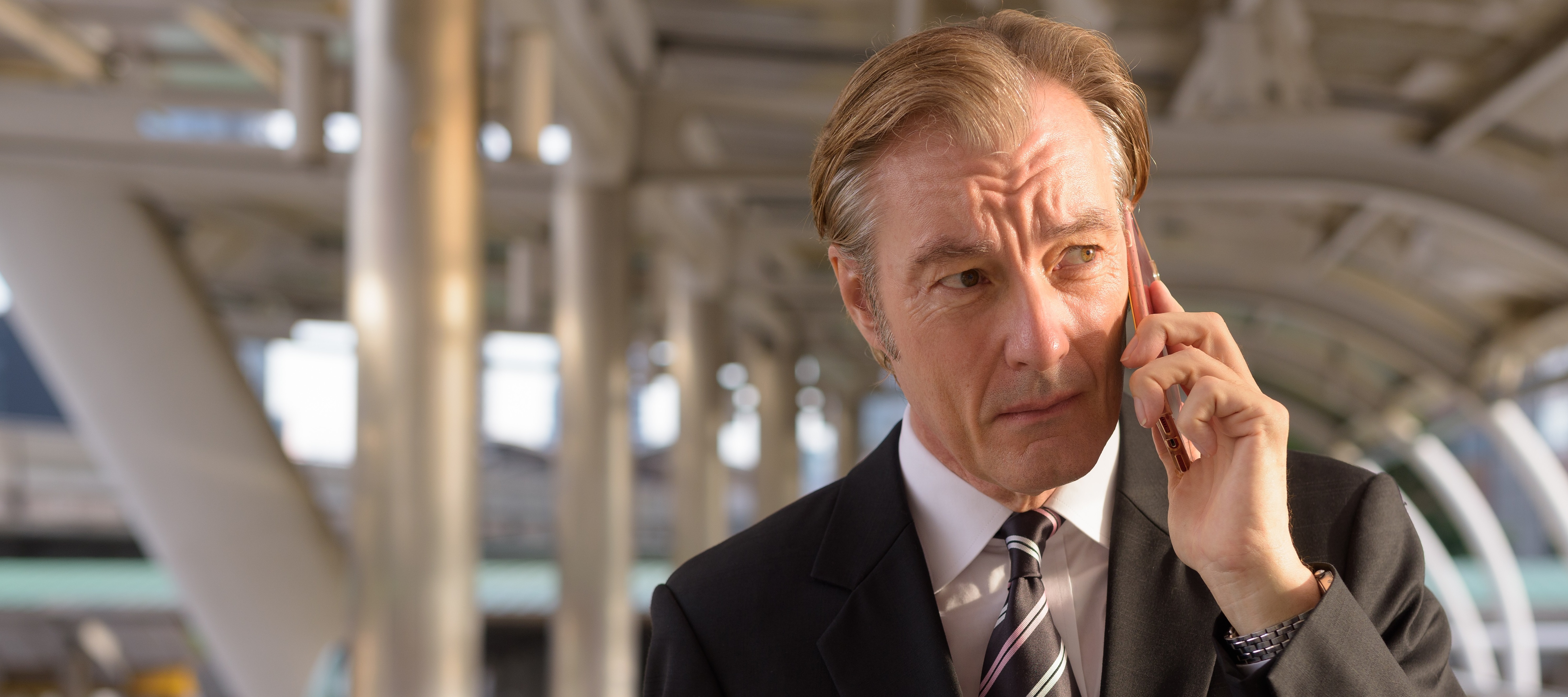 A serious-looking man with wearing a dark suit and tie holds a phone to his ear while standing outside.