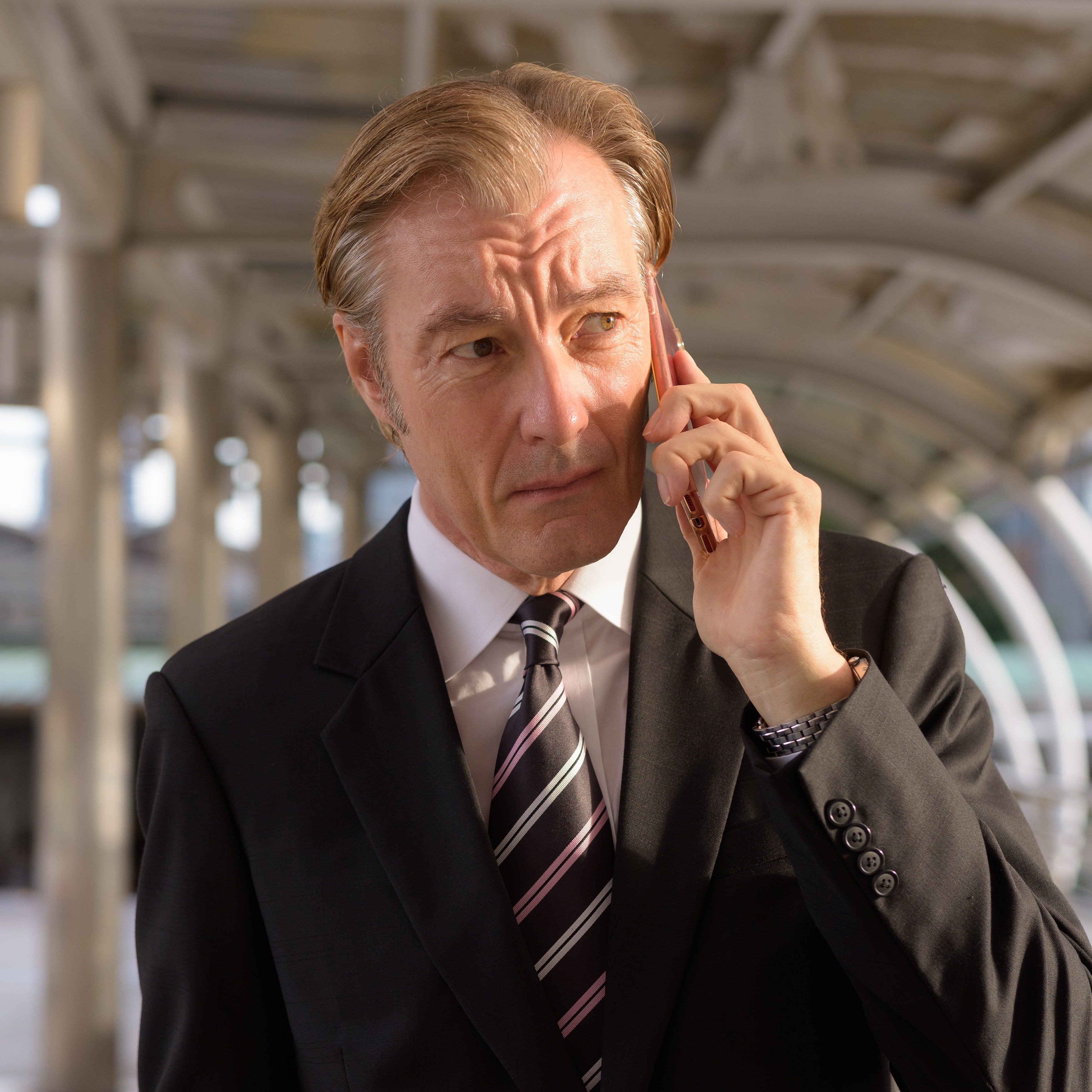 A serious-looking man with wearing a dark suit and tie holds a phone to his ear while standing outside.