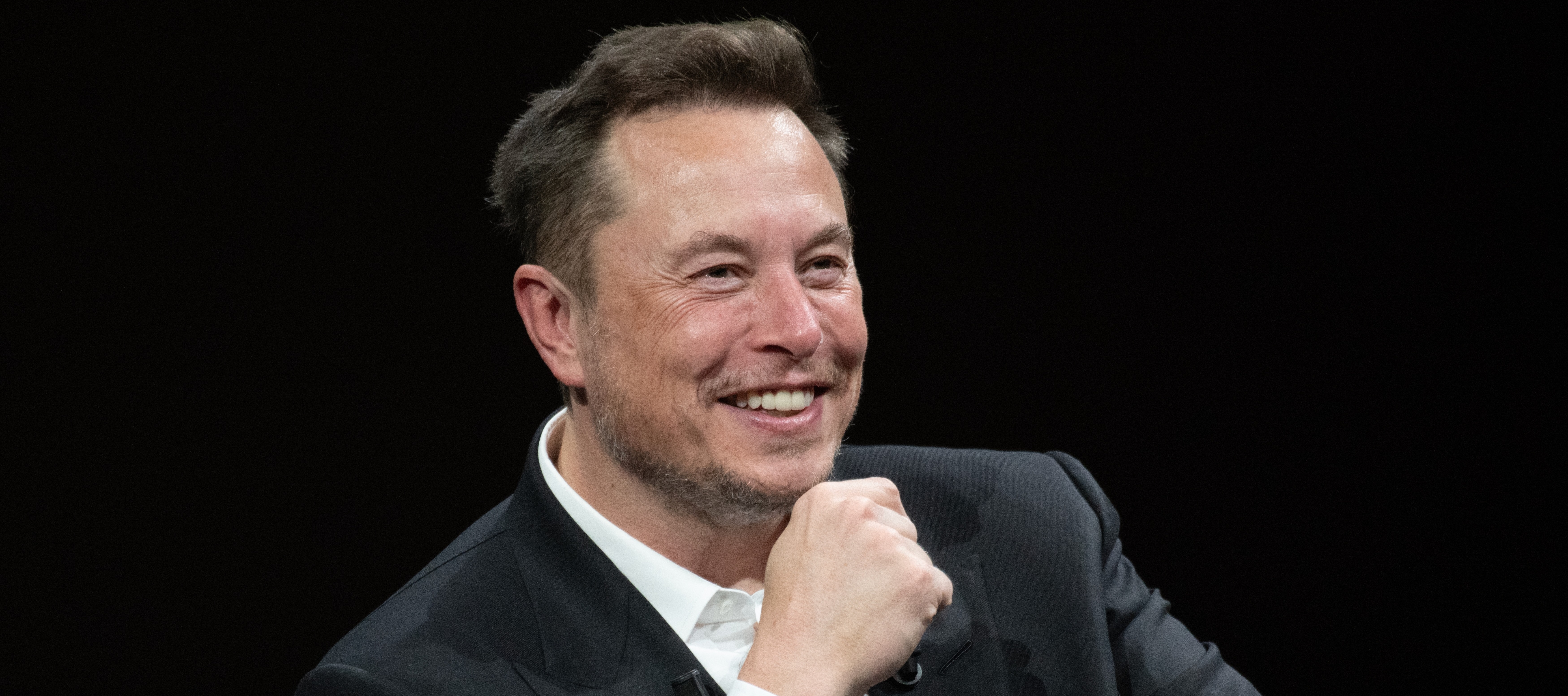 A close up of Elon Musk smiling broadly with a fist resting on his chin while he speaks on stage.