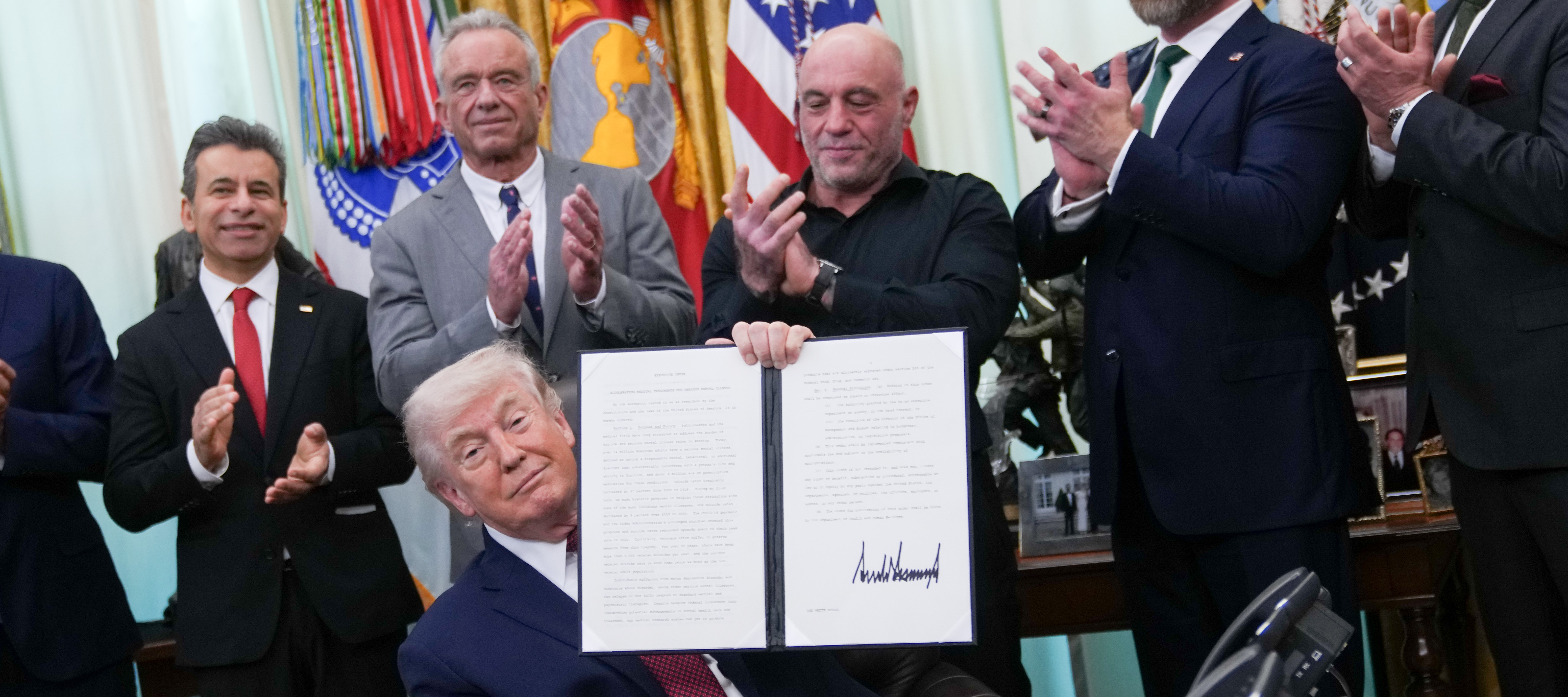 President Donald Trump holds up a signed executive order while Joe Rogan looks on from over his shoulder.