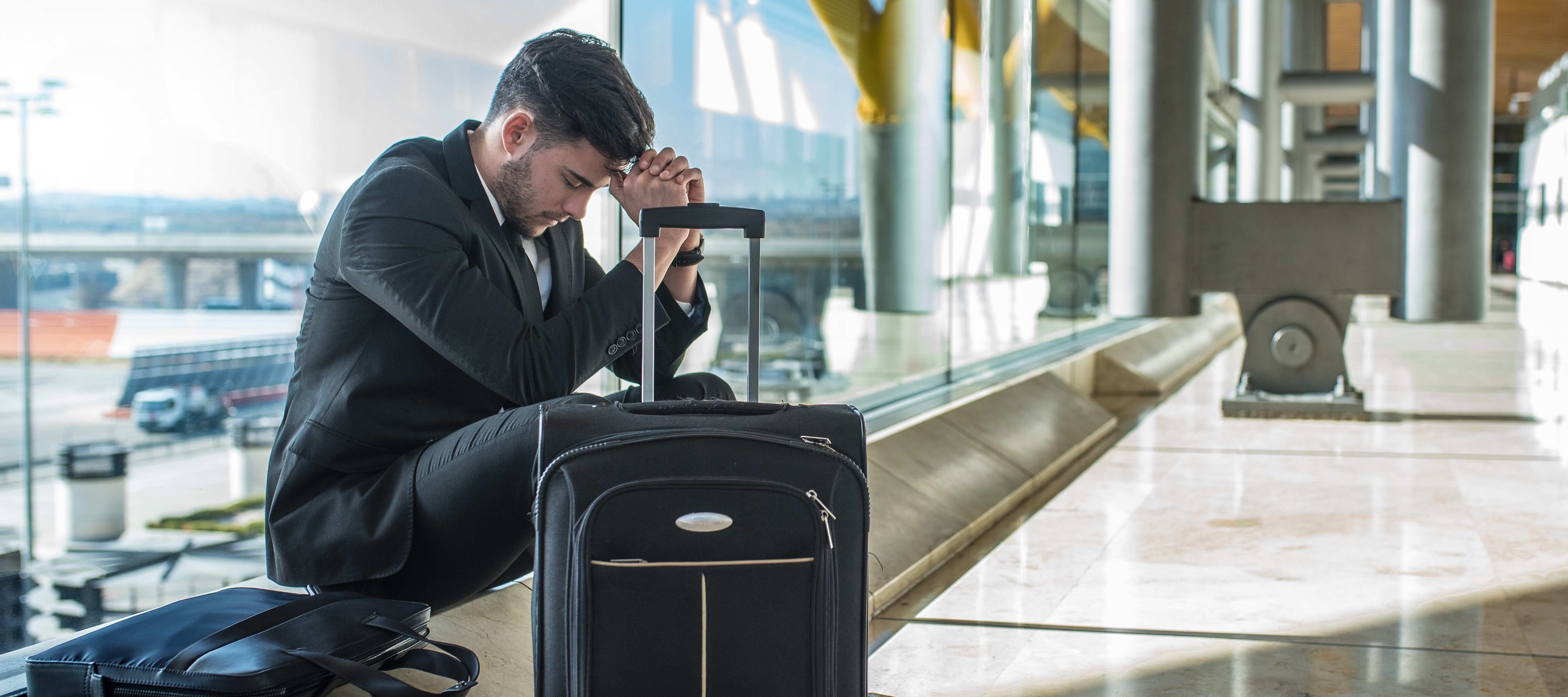 A smartly-dressed man sits in an airport hall with his head in his hands next to a suitcase and bag.