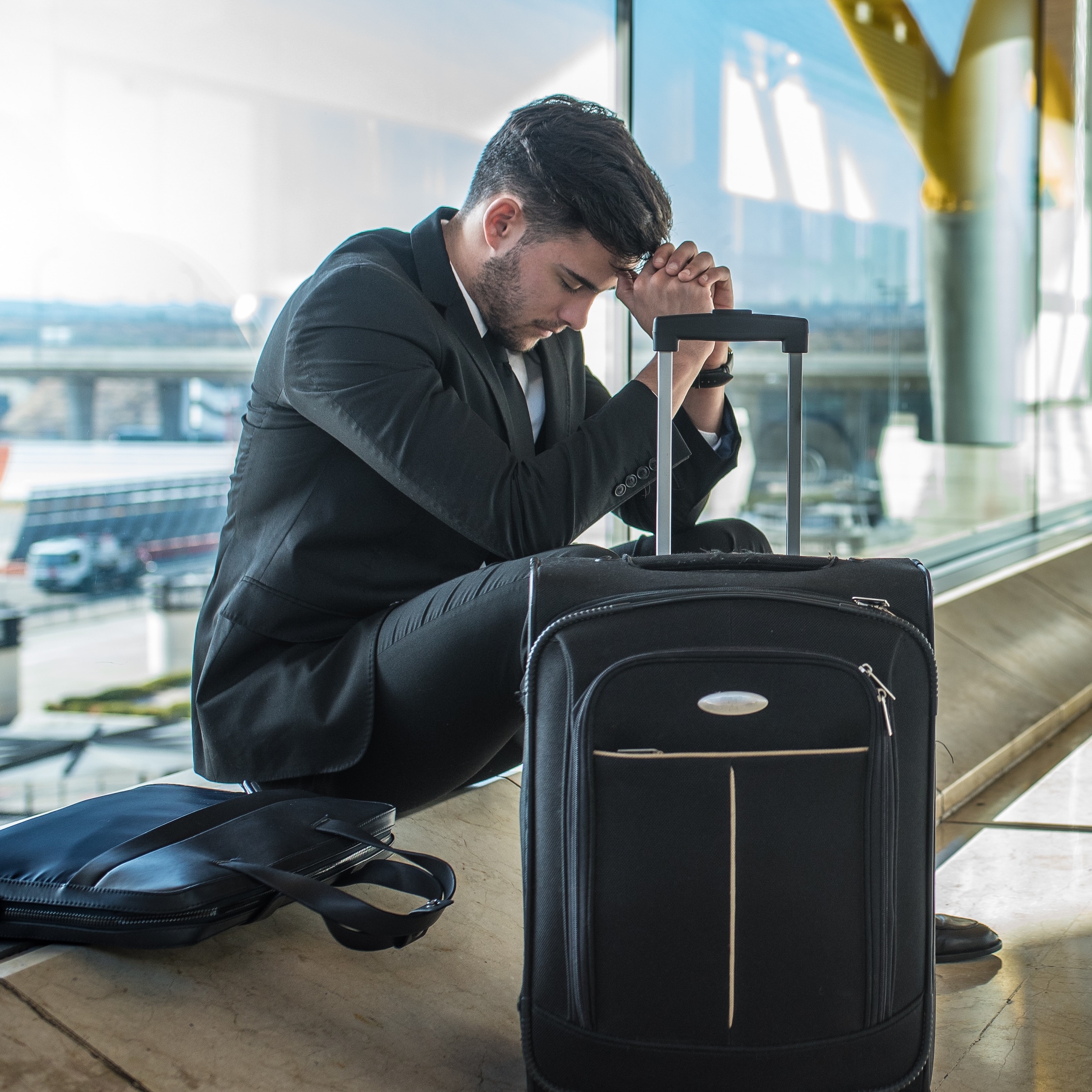 A smartly-dressed man sits in an airport hall with his head in his hands next to a suitcase and bag.