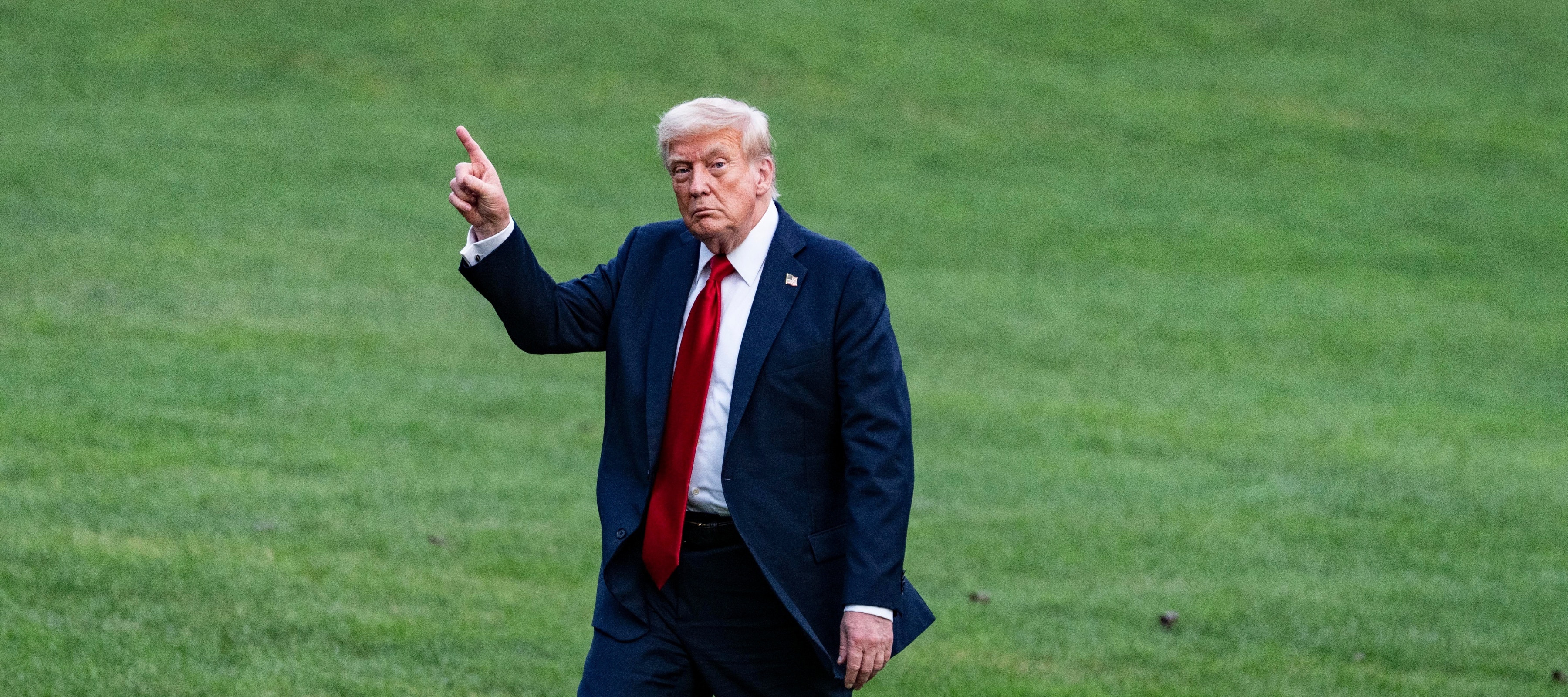 Donald Trump walking on a green lawn wearing a suit and pointing one finger in the air