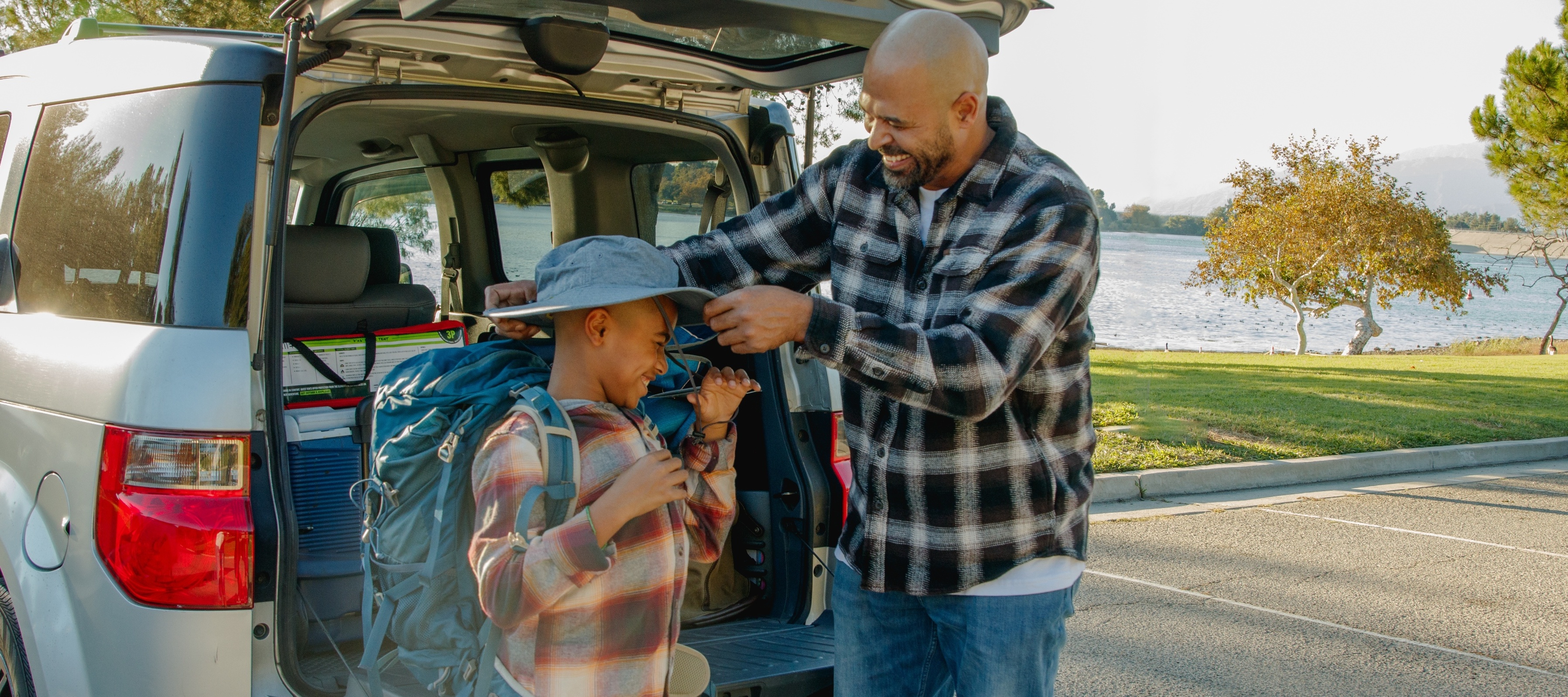 A father and son laugh as they unpack a car filled with camping gear.