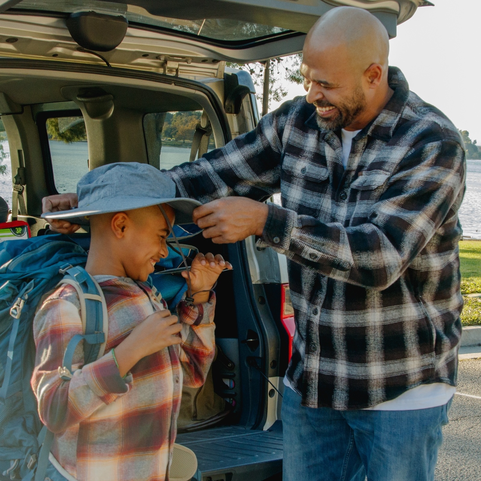 A father and son laugh as they unpack a car filled with camping gear.