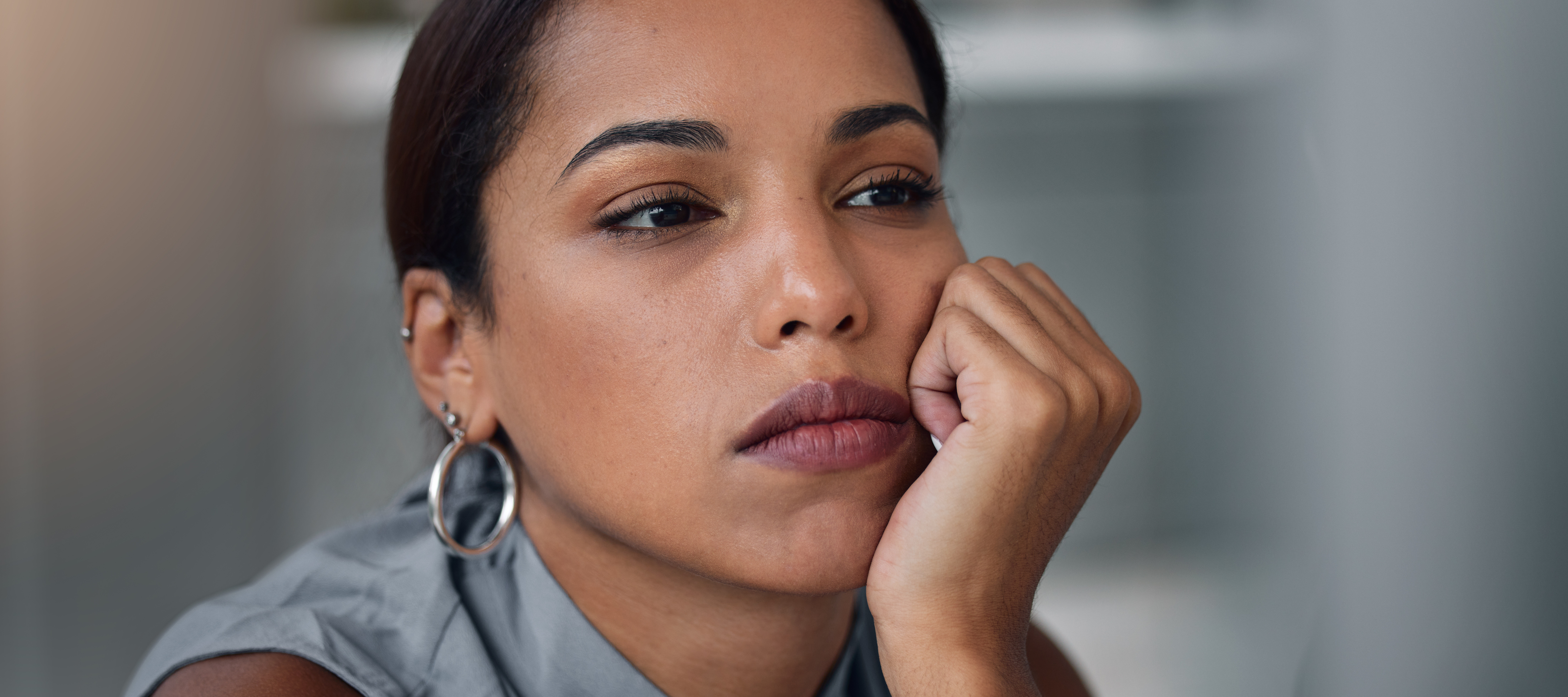 Woman with dark, straight hair, wearing a gray top, rests her head on her hand with a pensive expression.