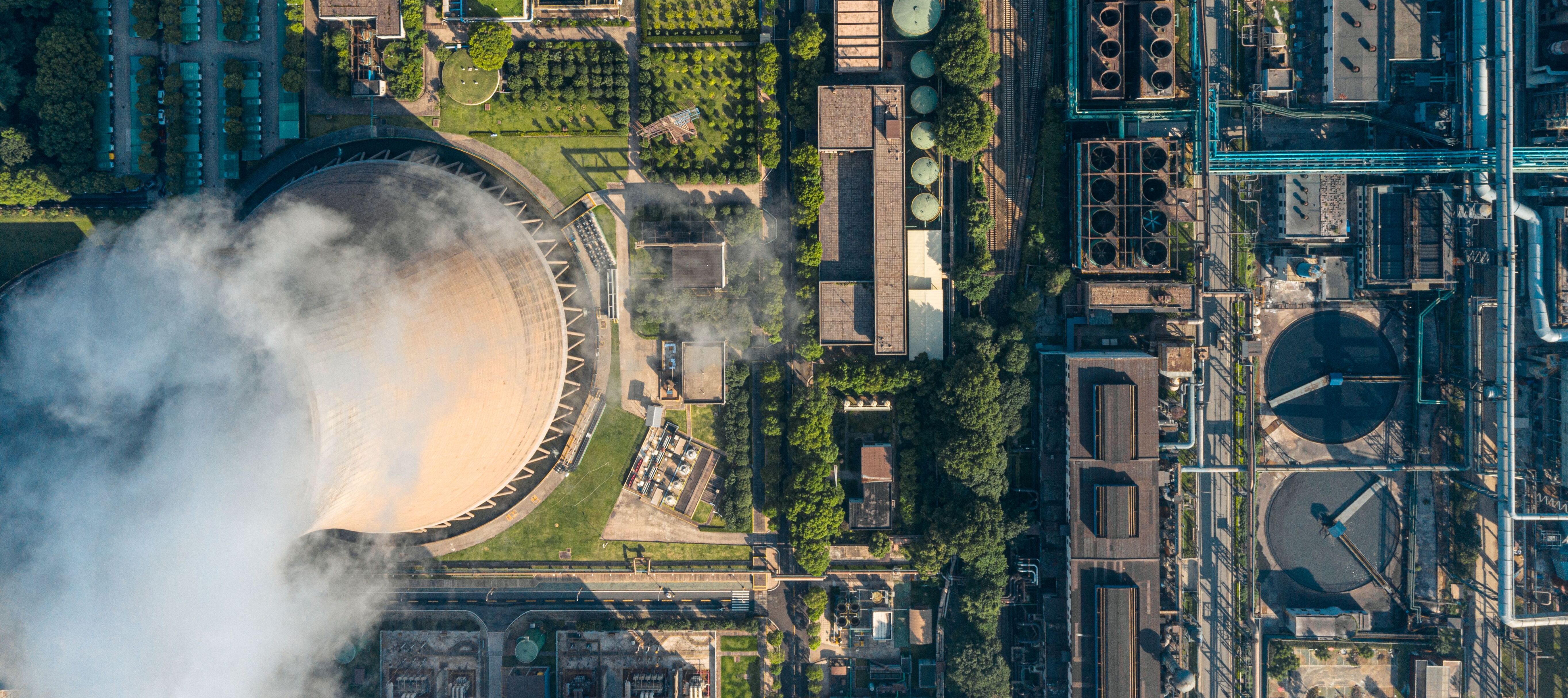 An aerial view showcases green spaces, trees, and manicured landscaping sit beside industrial structures like a beige cooling tower with escaping white clouds.