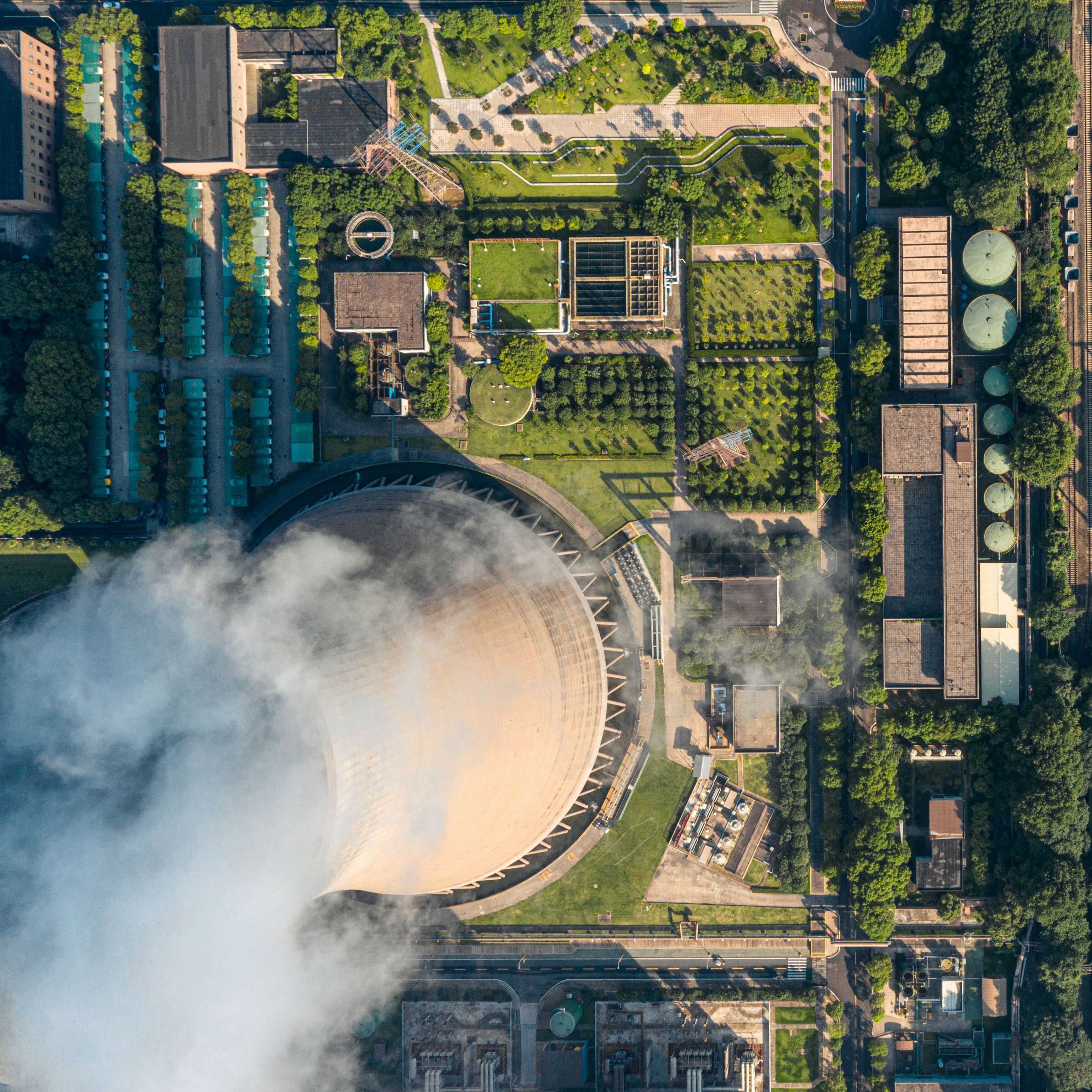 An aerial view showcases green spaces, trees, and manicured landscaping sit beside industrial structures like a beige cooling tower with escaping white clouds.