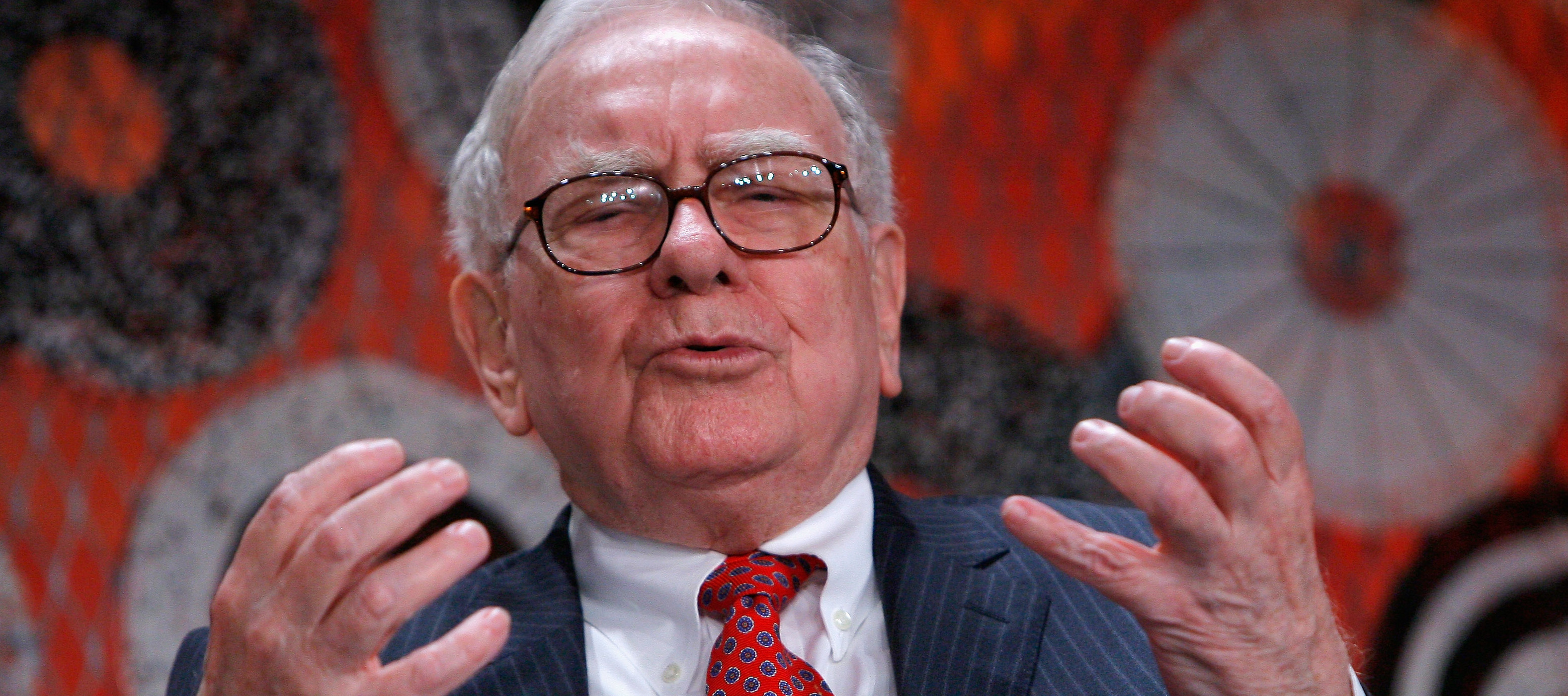 Warren Buffett holds up both hands, grimacing against a backdrop of white, black and red concentric circles..
