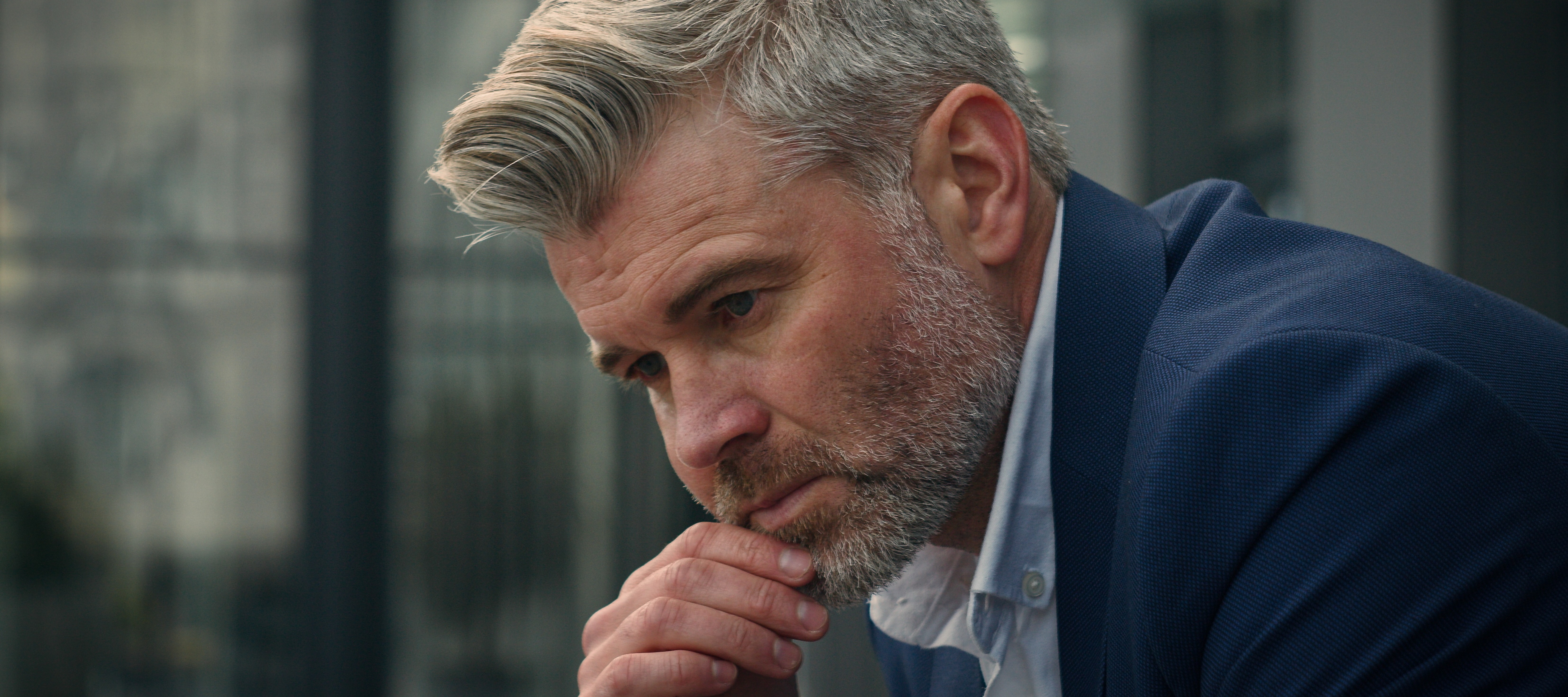 adult man in a suit, his brow furrowed in apparent thought. His hair is light and his facial hair is short and groomed. He is resting his chin on his hand.