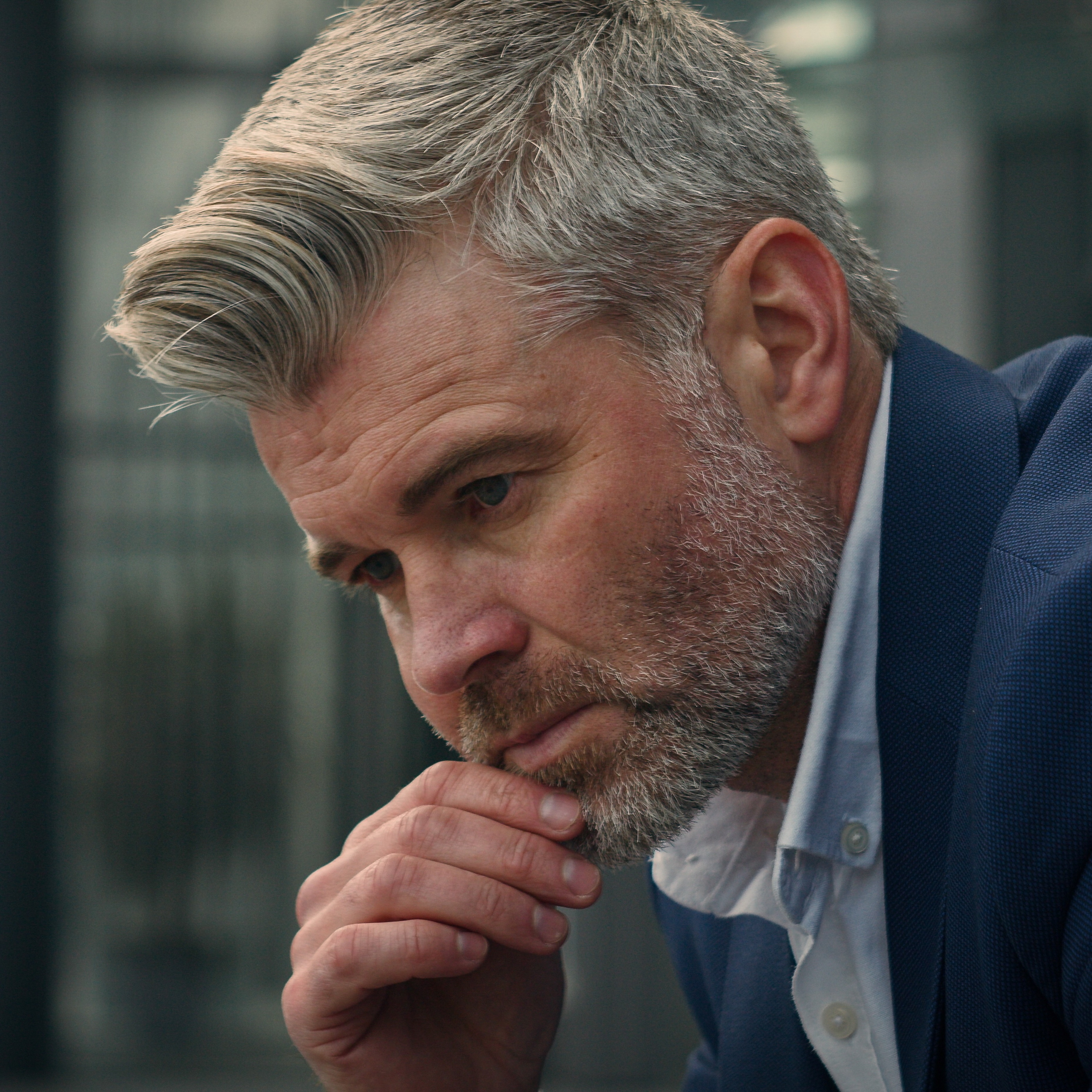 adult man in a suit, his brow furrowed in apparent thought. His hair is light and his facial hair is short and groomed. He is resting his chin on his hand.