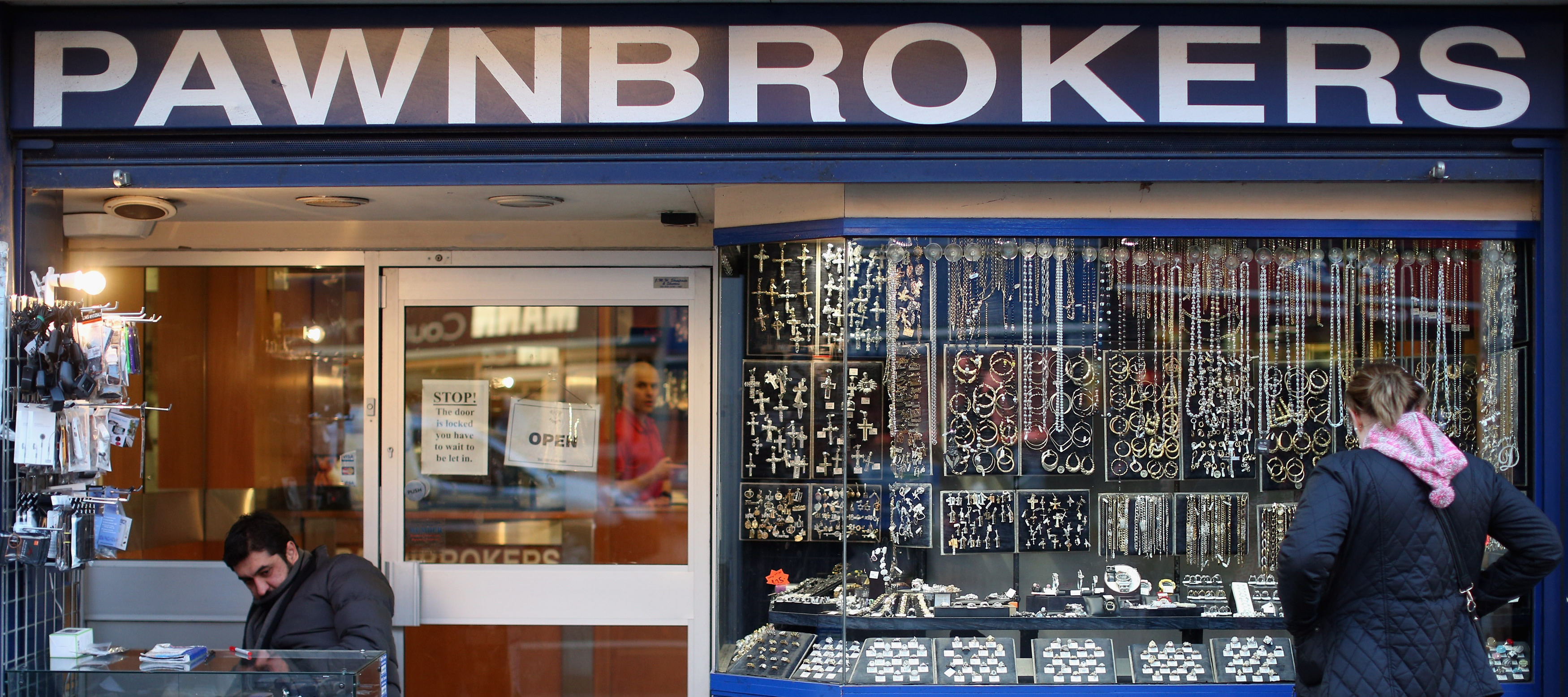 A woman looks into a pawnbroker shop window that is filled with watches and jewelry on a city street.