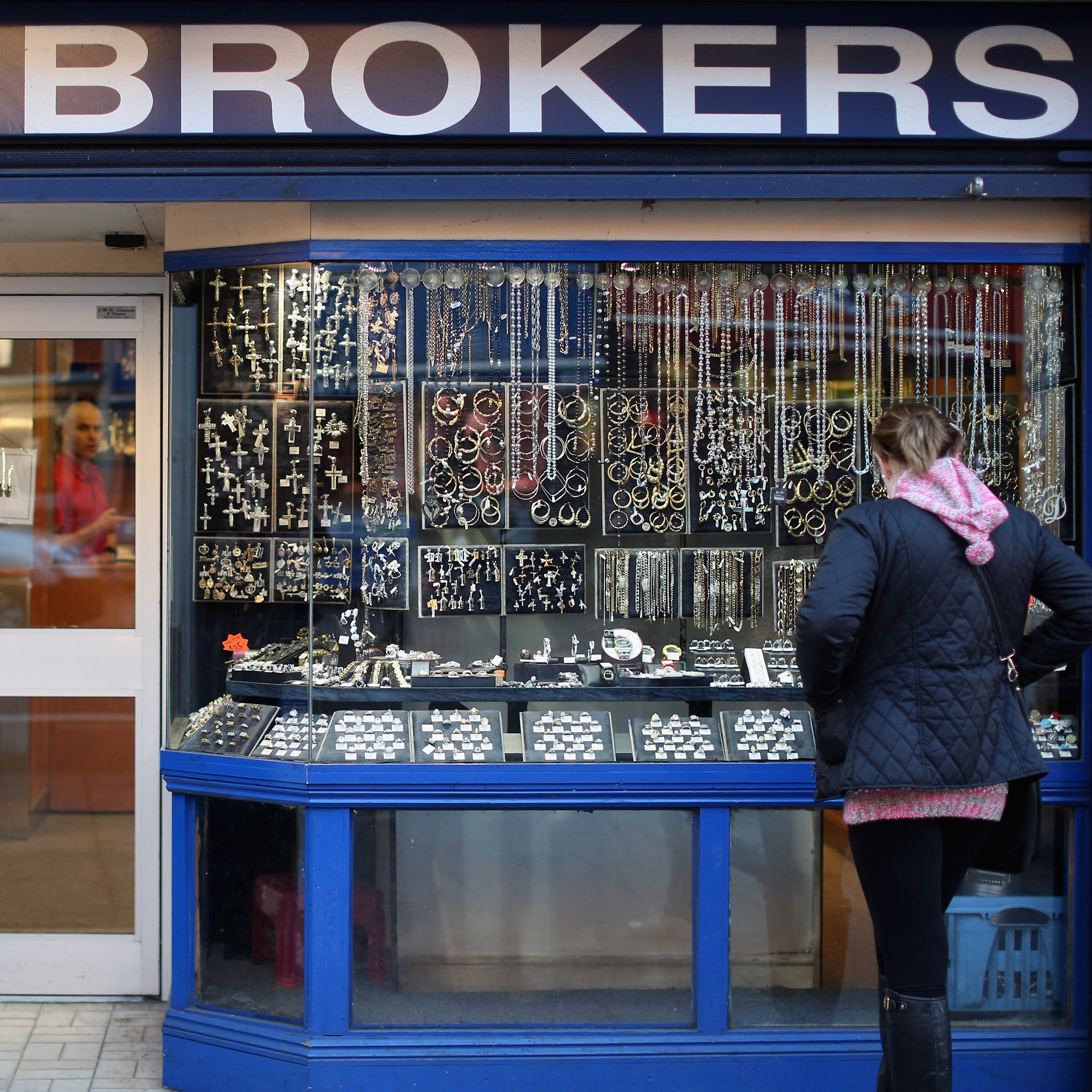 A woman looks into a pawnbroker shop window that is filled with watches and jewelry on a city street.